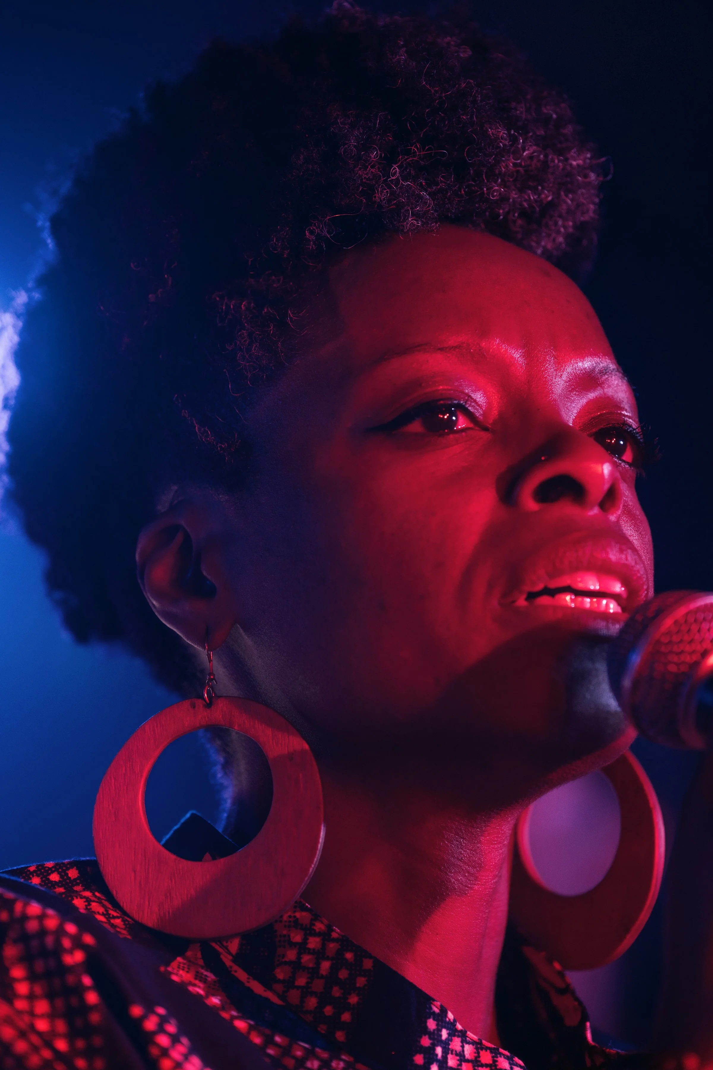 Retrato cercano de una mujer con cabello rizado y grandes aretes circulares. Luz rosa y azul en un entorno de escenario, con micrófono.