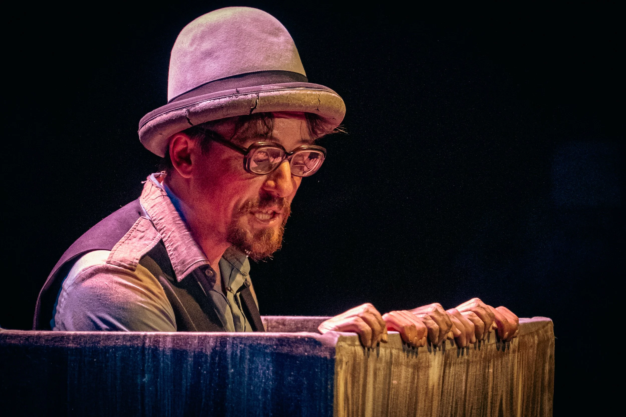 Un hombre con gafas, sombrero de fieltro y ropa casual toca un piano en un escenario oscuro.