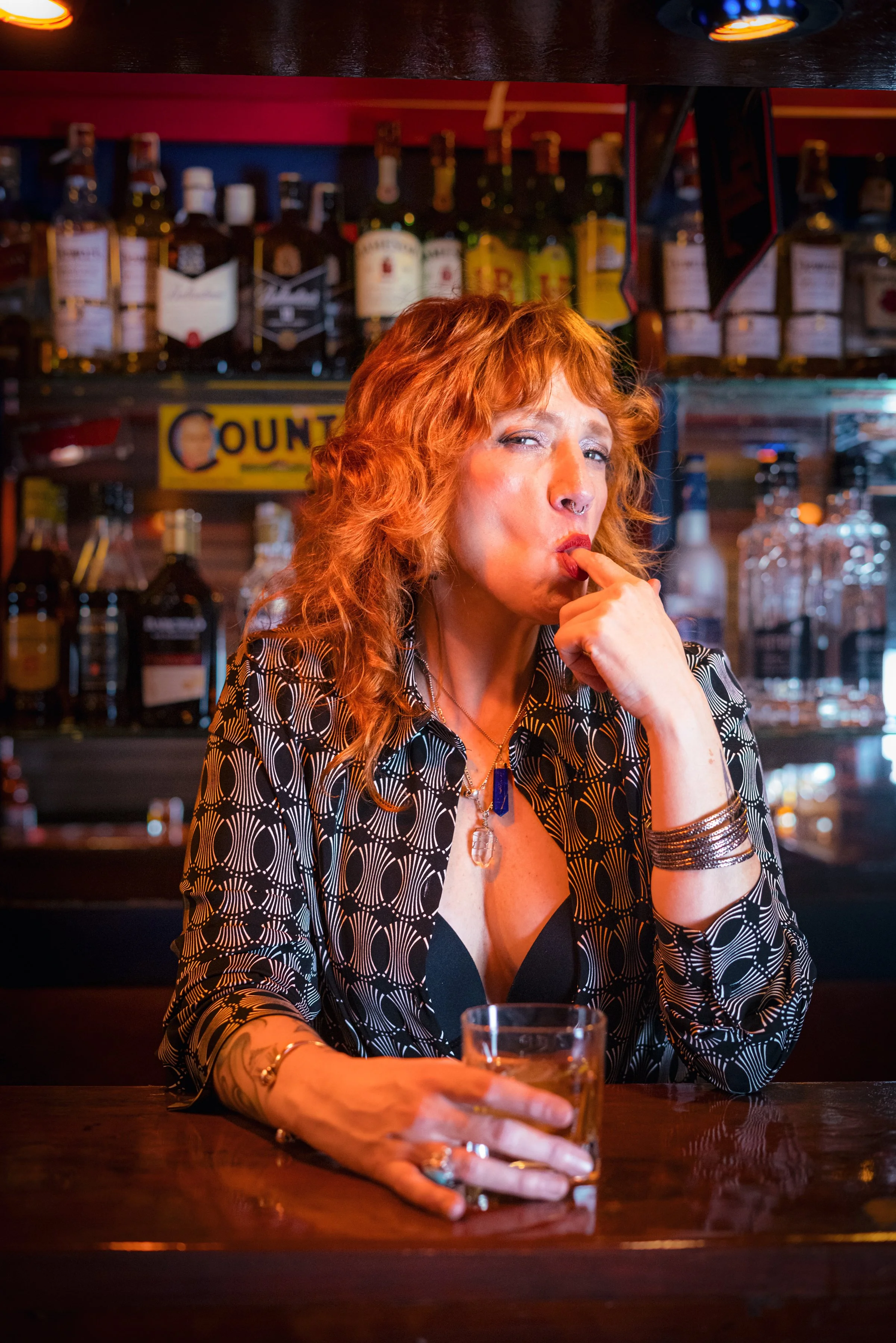 Mujer pelirroja en bar, con vaso en mano, mordiendo su dedo mientras mira a la cámara.