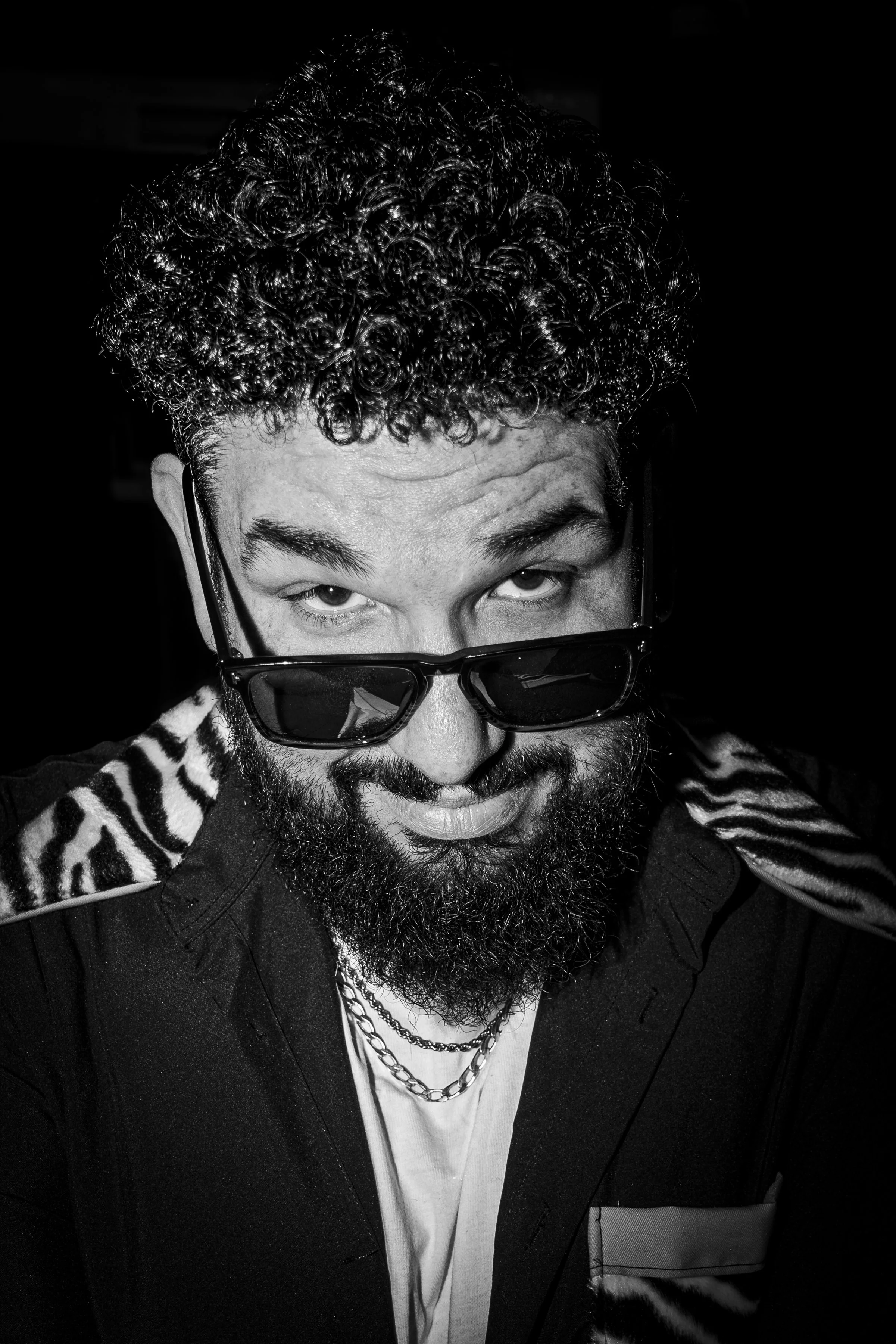 Retrato en blanco y negro de un hombre con cabello rizado y barba, usando gafas de sol y una camiseta blanca con chaqueta negra, con joyas, en una pose con expresión coqueta y mirada elevada.