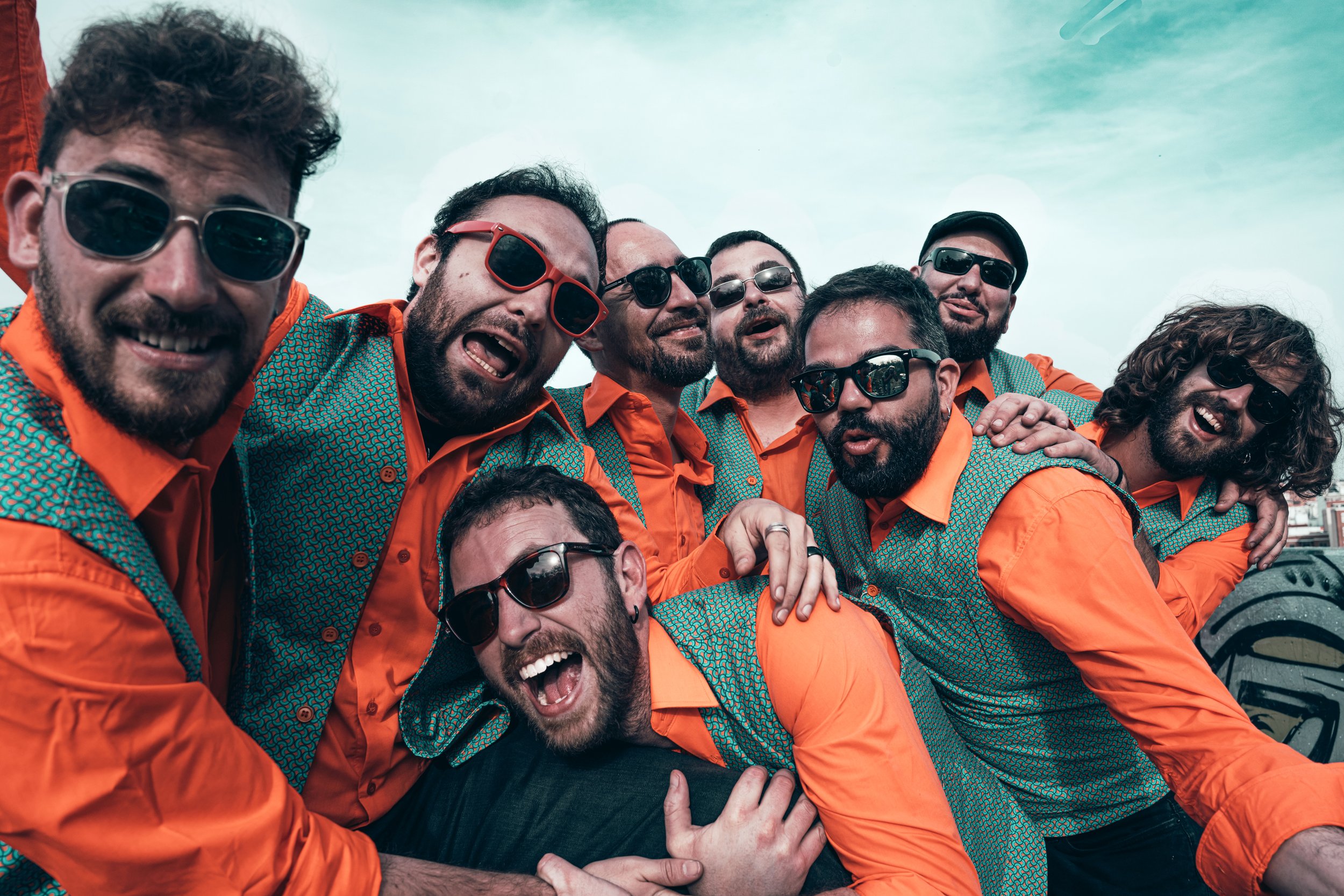 Un grupo de hombres sonriendo y abrazándose en un ambiente exterior, vestidos con chaquetas de color verde con patrón y camisa naranja, todos con gafas de sol.