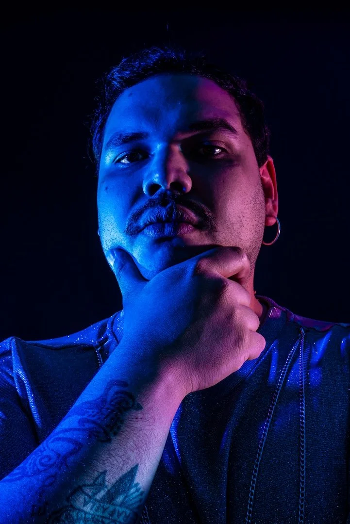 Retrato de un hombre con barba y barba rizada, con tatuajes en el brazo, usando auriculares y camiseta de color oscuro, en un fondo negro con iluminación azul y púrpura.
