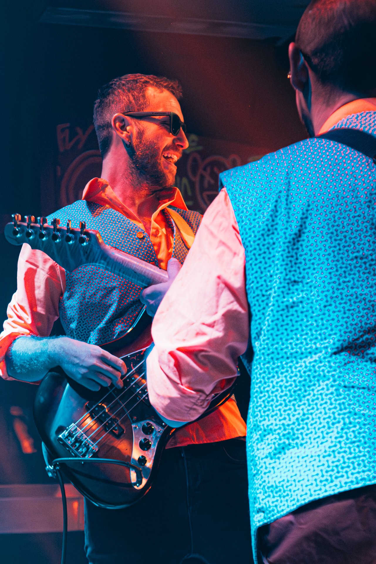 Hombre tocando guitarra eléctrica y sonriendo, usando gafas de sol y camisa colorida, en un escenario con iluminación de colores cálidos.