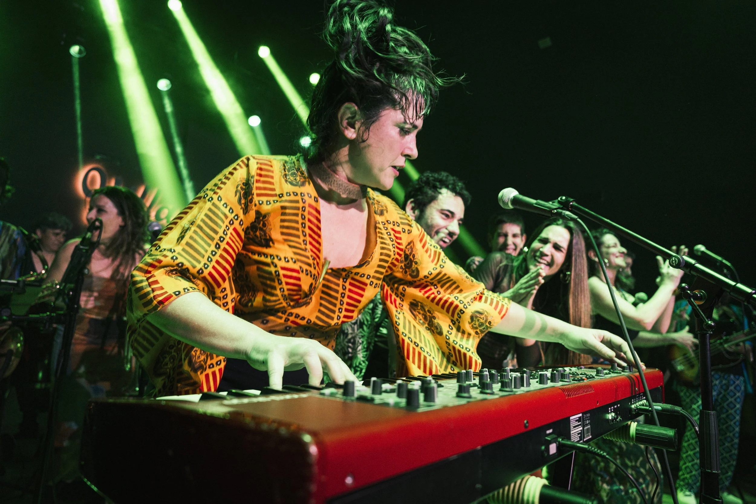 Mujer tocando un teclado en un concierto con luces verdes y varias personas sonriendo y disfrutando la música en el fondo.