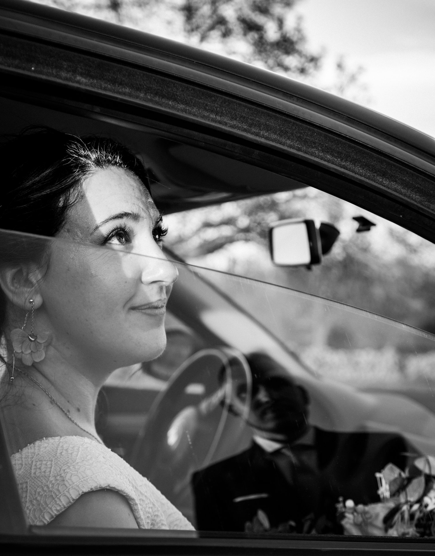Mujer con vestido encendiendo mientras está en el asiento del conductor, con un hombre vestido en uniforme militar o de ceremonia en el asiento del copiloto, en un coche. La imagen es en blanco y negro.