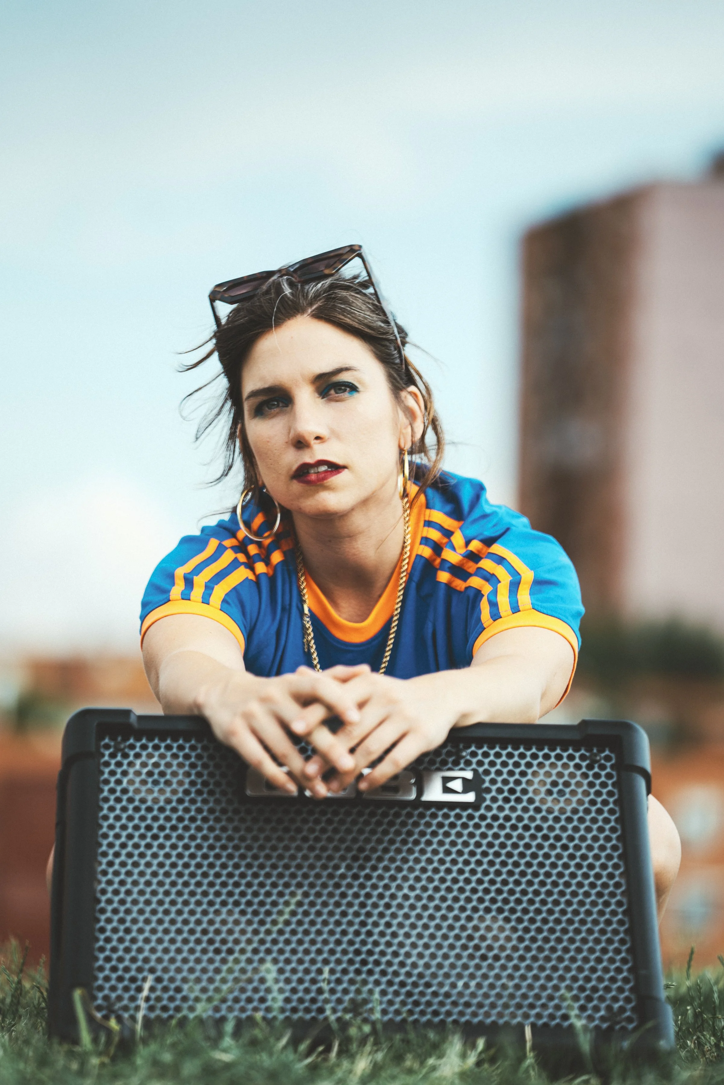 Joven mujer con maquillaje oscuro, camiseta azul con franjas amarillas, lentes en la cabeza, collares y pendientes, sentado sobre césped con altavoz portátil.