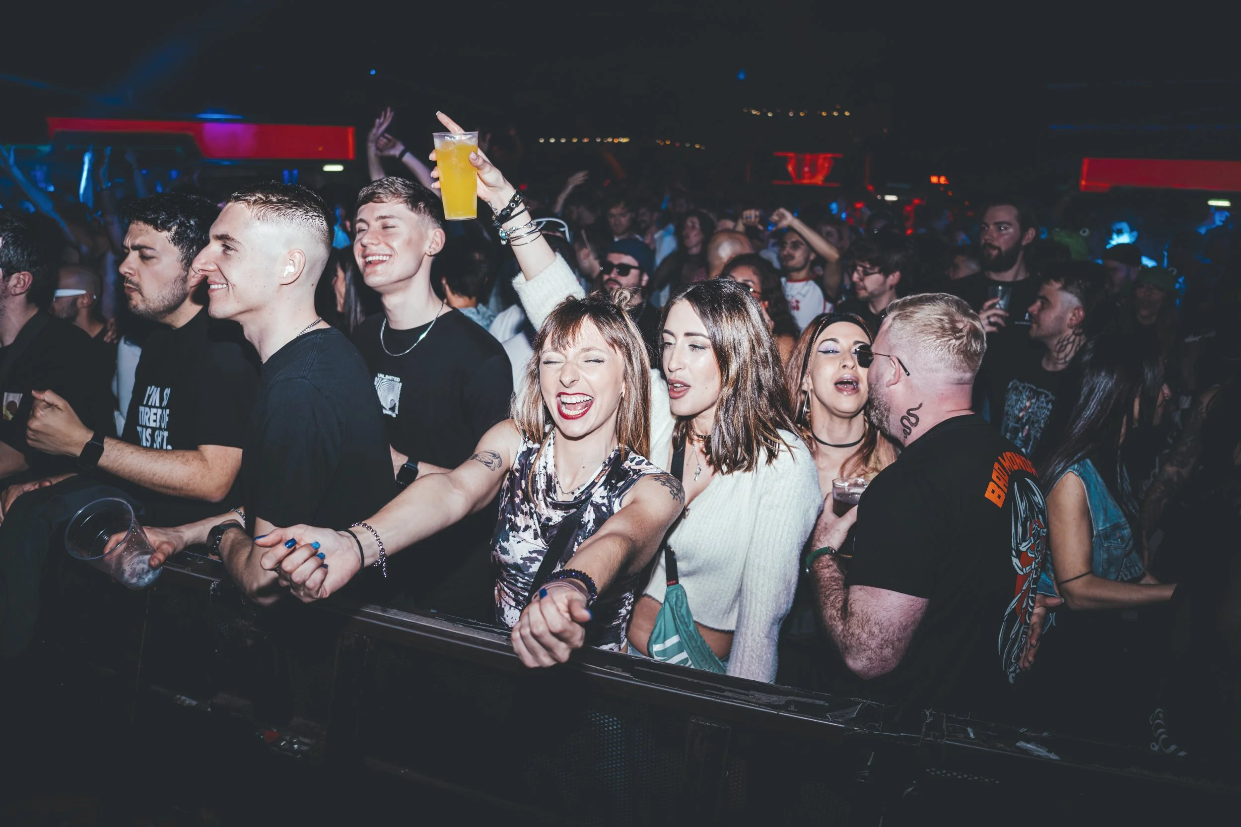 Grupo de personas disfrutando en un concierto, algunos bailando, riendo y con bebidas en mano en un ambiente oscuro con luces de colores.