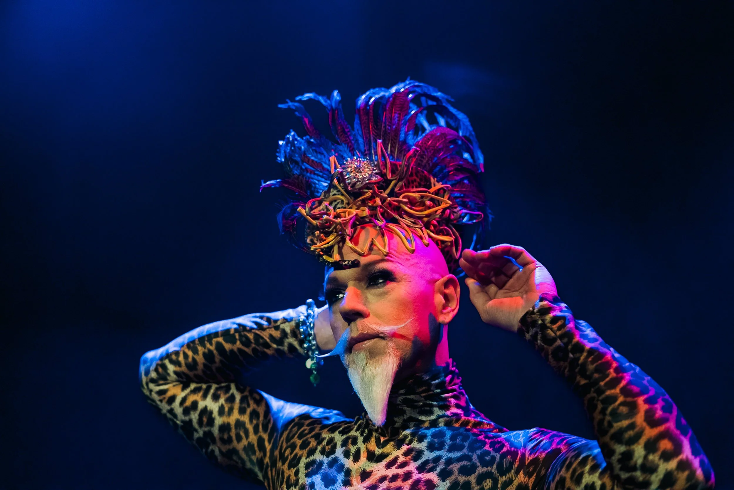 Persona con maquillaje llamativo, pelo alto decorativo con plumas de colores, ropa de leopardo, en un escenario con luz azul y rosa.