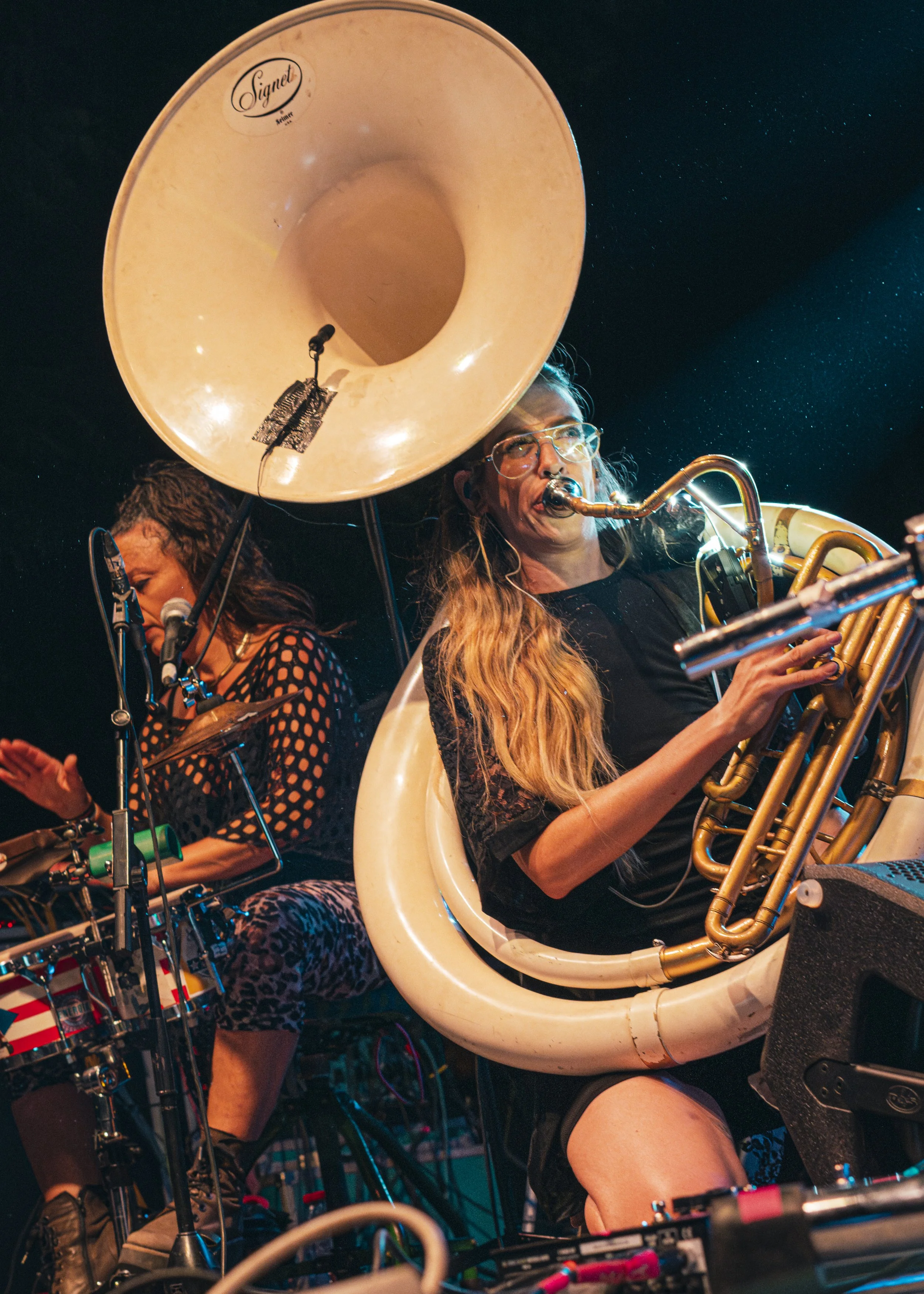 Dos mujeres tocando instrumentos musicales en un escenario, una con un saxofón y la otra con una batería, con un fondo oscuro y luces de escenario.