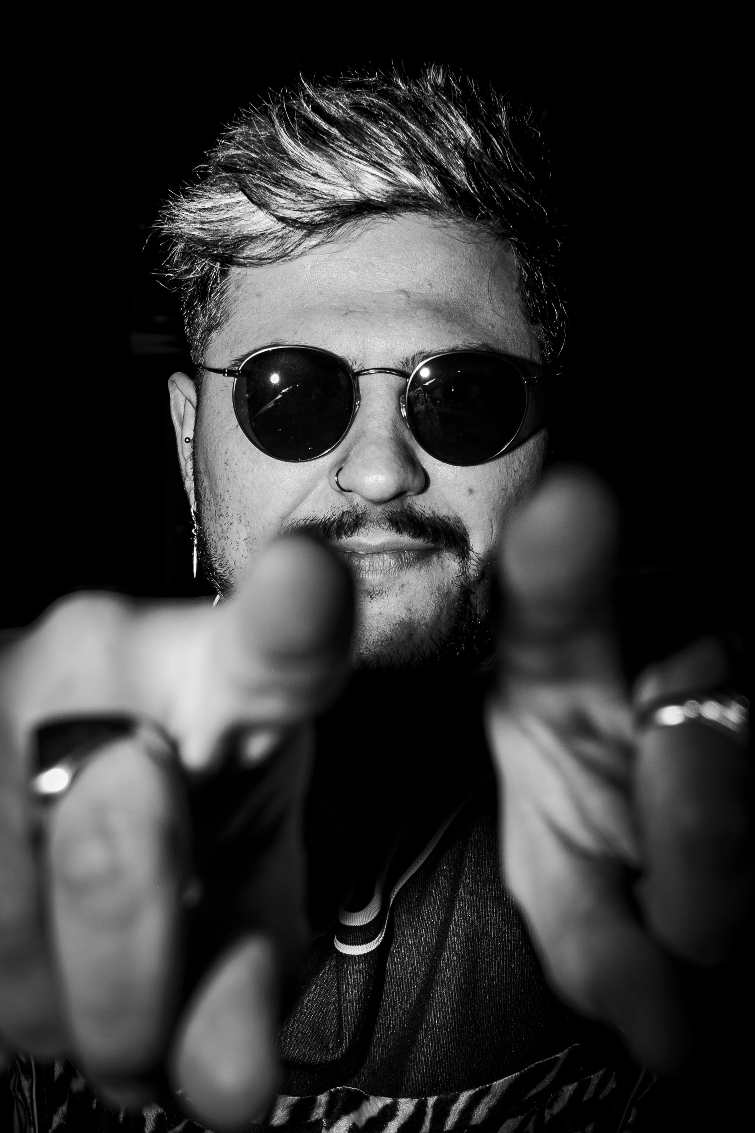 Retrato en blanco y negro de un hombre con gafas de sol redondas, barba, bigote, y una expresión confiada, mirando directamente a la cámara con las manos en primer plano.