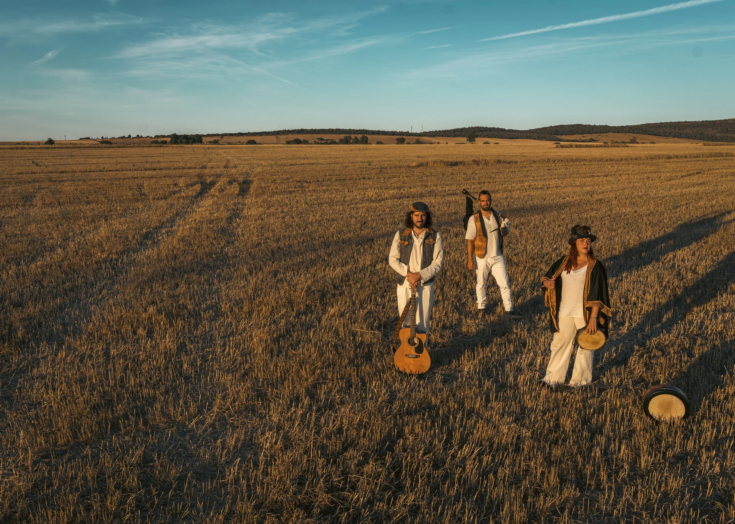 Tres personas en un campo dorado con instrumentos musicales y vestimentas de estilo folclórico, durante el atardecer