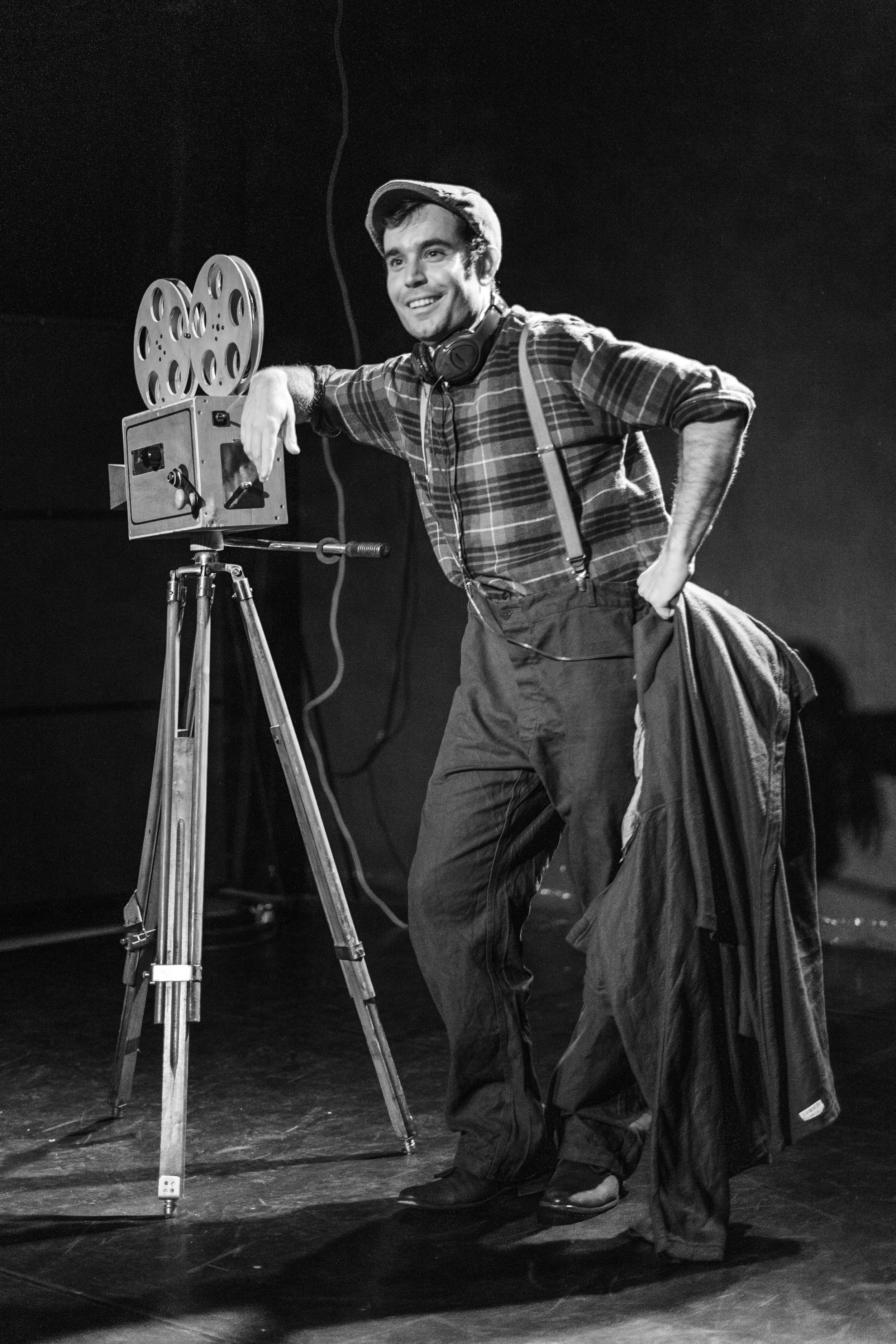 Hombre con camisa a cuadros, sombrero y auriculares, posando junto a una cámara de cine antigua en un escenario oscuro, sonriendo.