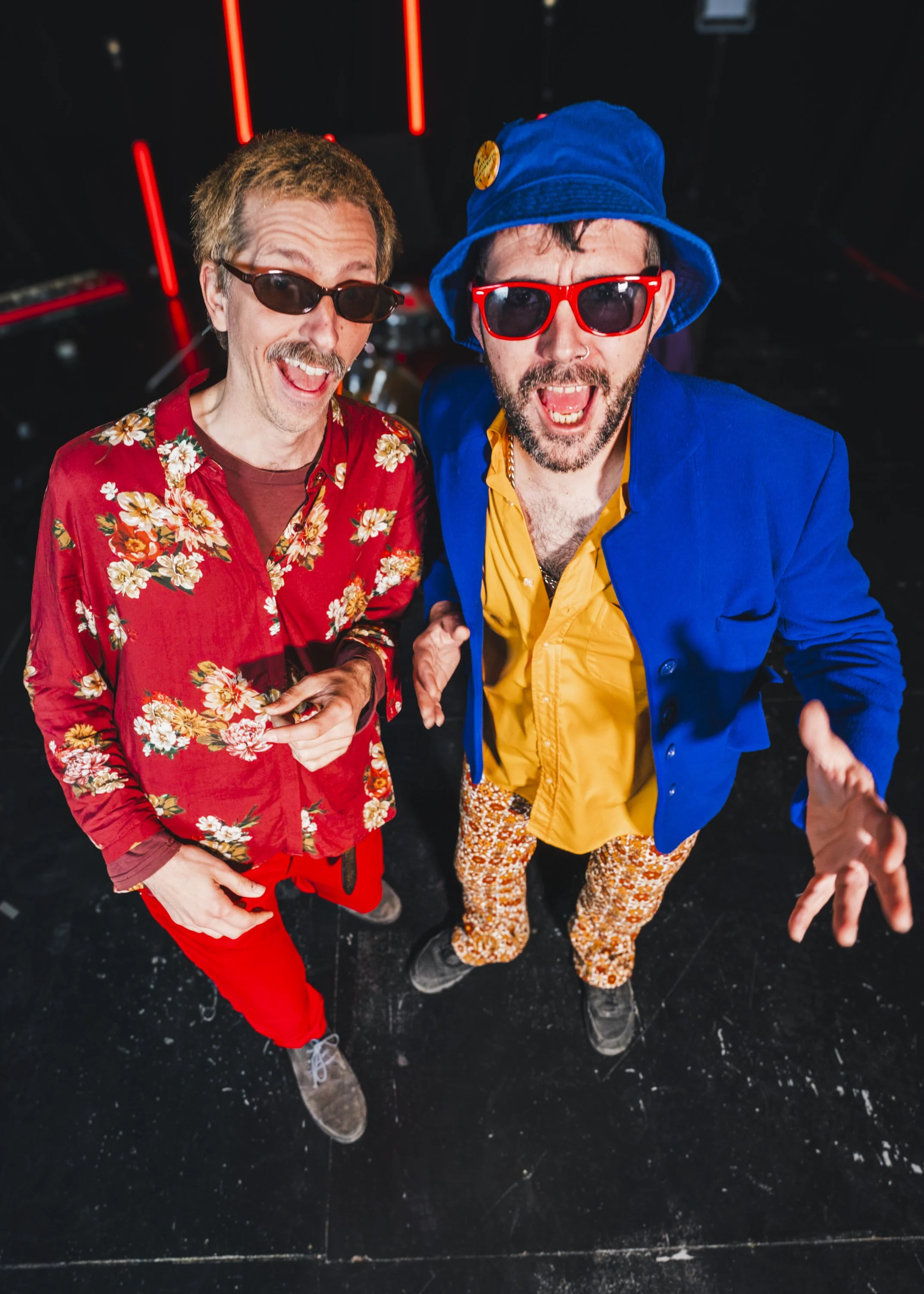 Dos hombres con ropa colorida, gafas y accesorios, sonriendo y posando para la cámara en un ambiente con iluminación de neón y suelo negro