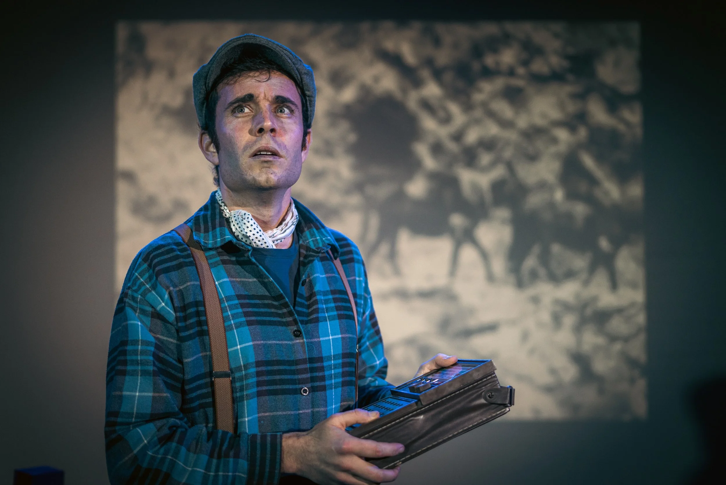 Actuación teatral de un hombre con vestimenta clásica, sosteniendo un libro o guion en un escenario con fondo de proyección.