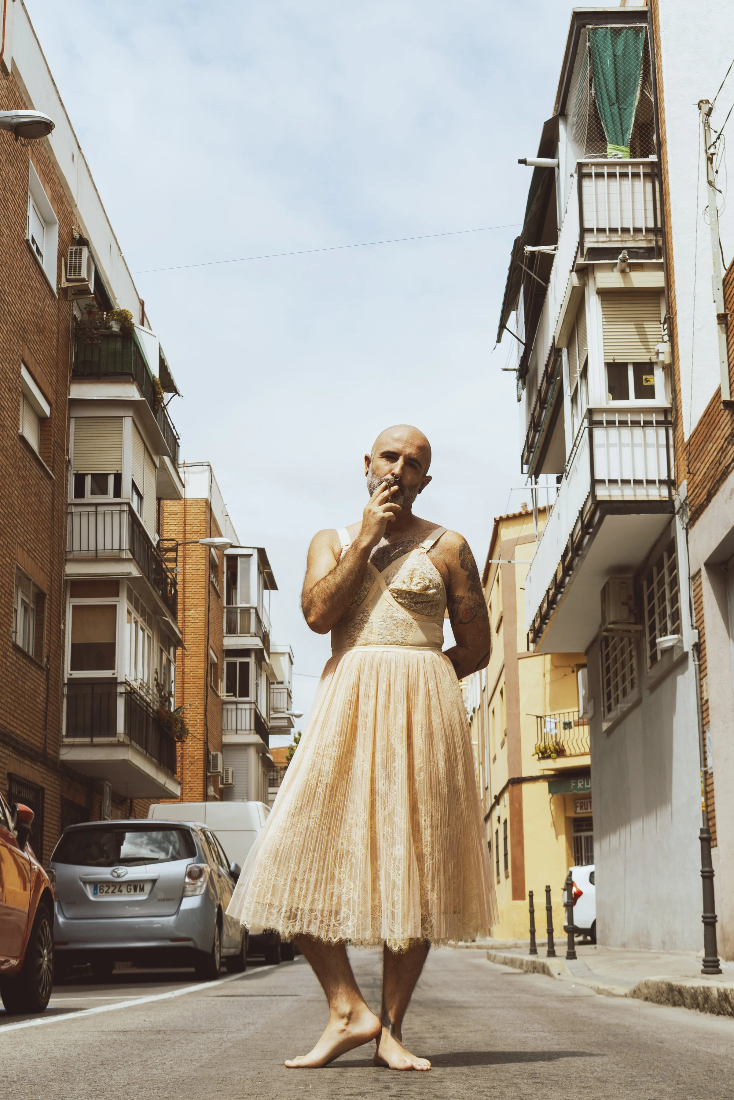 Hombre con tatuajes vestido con un vestido de tul color durazno, caminando sin zapatos en la calle de una ciudad con edificios de apartamentos de color ladrillo y balcones.