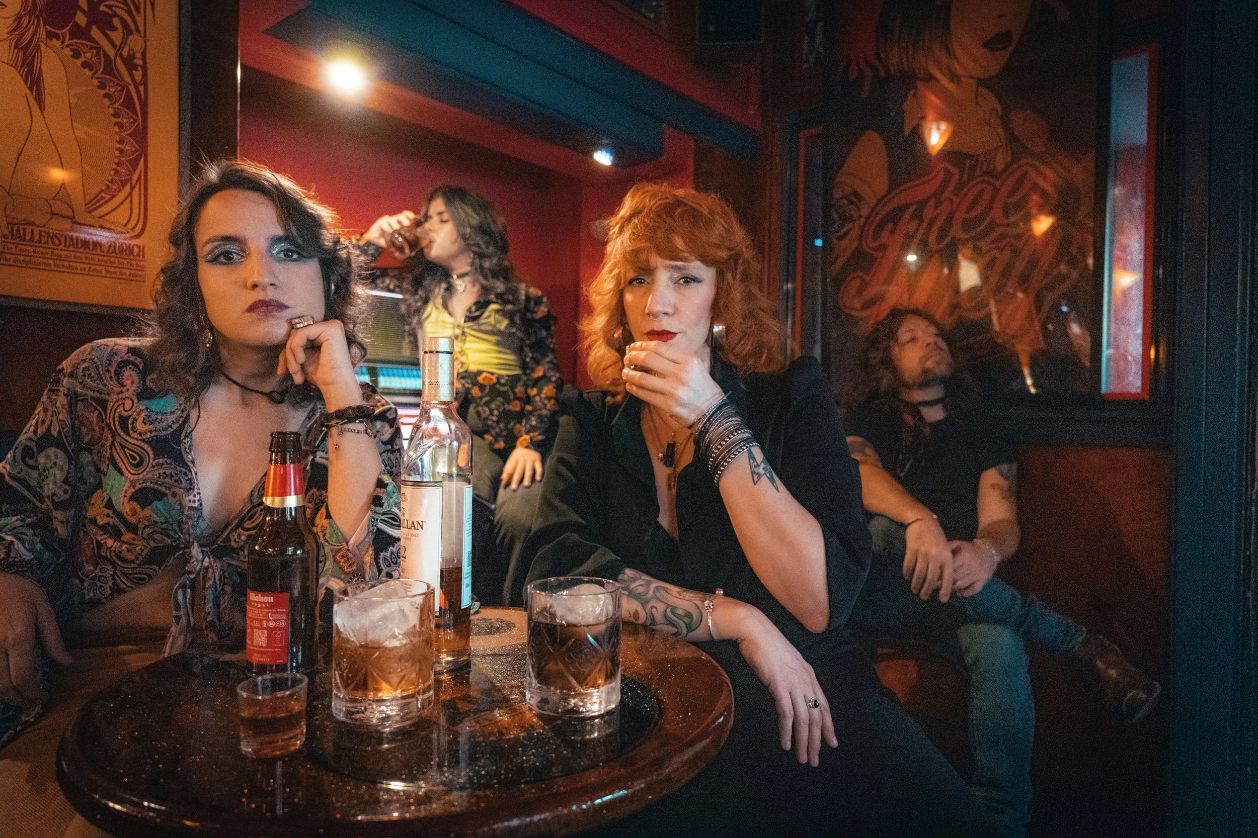 Cuatro personas en un bar, dos mujeres en primer plano, una con cabello rizado y otro con cabello encanecido, sentadas en una mesa con varias bebidas. En el fondo, otras dos personas, una tomando una bebida y otra recargada en la pared, en un ambient