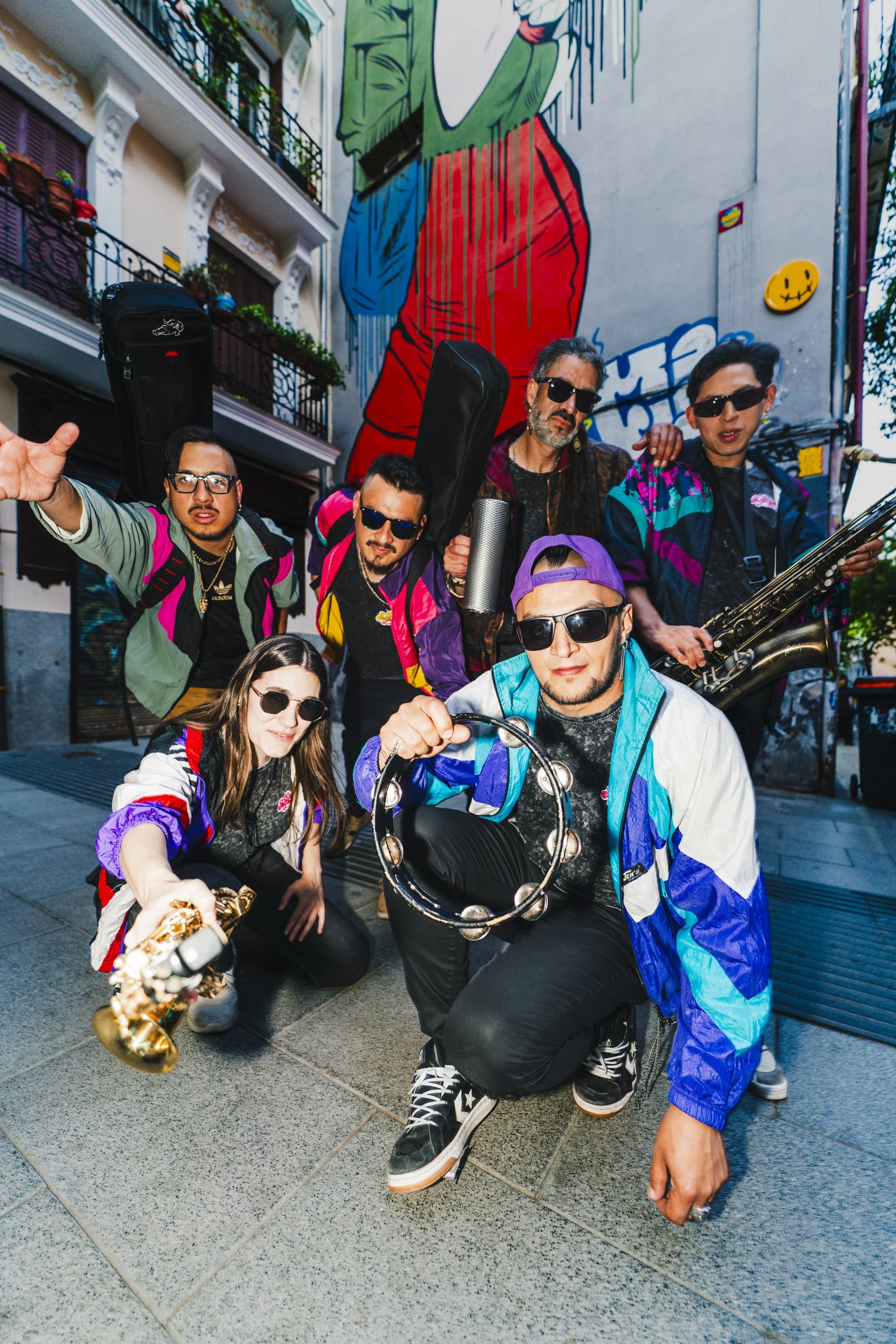 Grupo de personas jóvenes posando en una calle con graffiti en la pared trasera, algunos con instrumentos musicales, vistiendo ropa colorida y lentes oscuros.