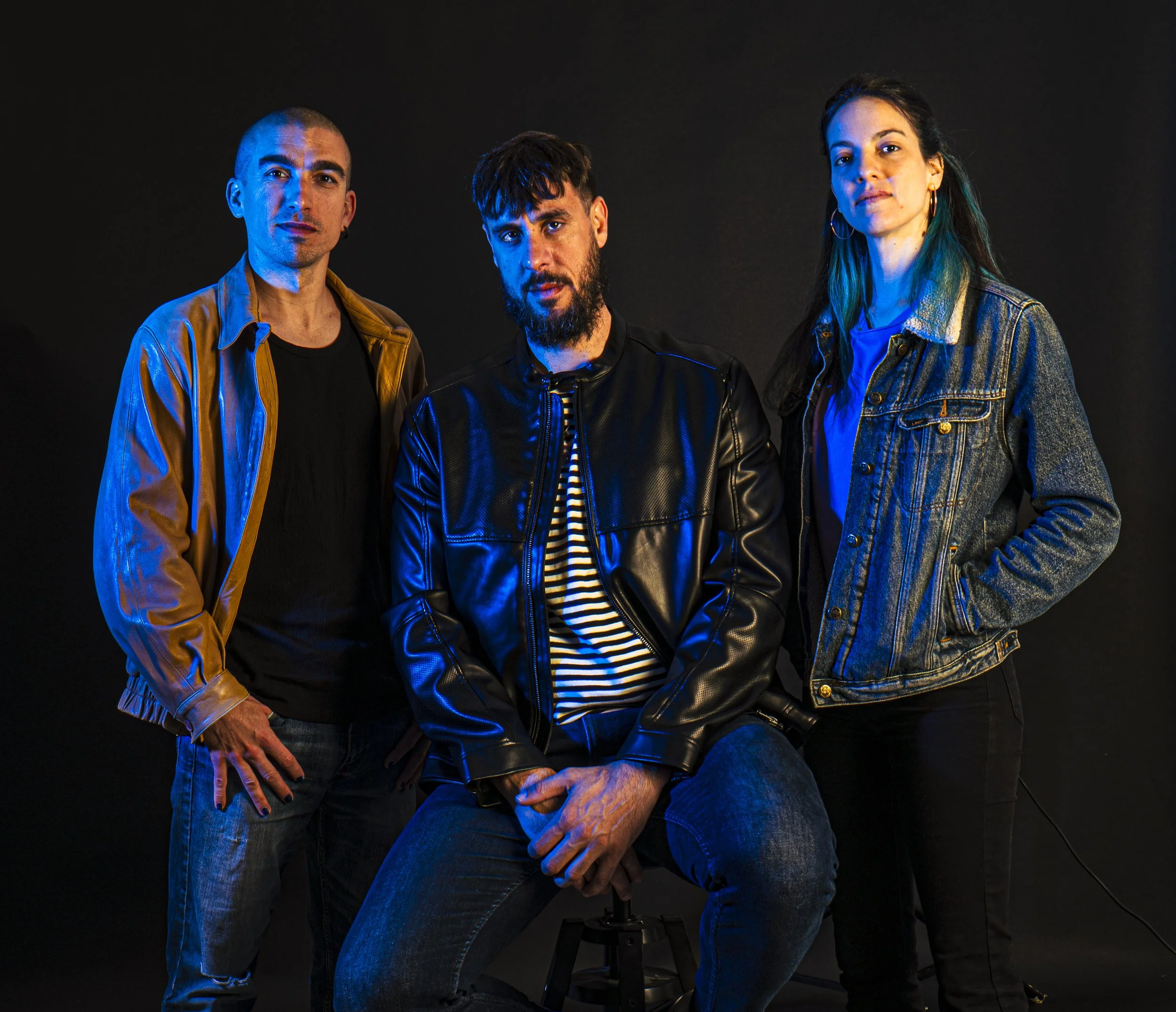 Tres personas en un fondo negro, con iluminación azul, en actitud relajada. Un hombre de cabello corto y barba en el centro, usando chaqueta de cuero negra y camiseta a rayas, sentado en un taburete. A la izquierda, otro hombre con chaqueta de color 