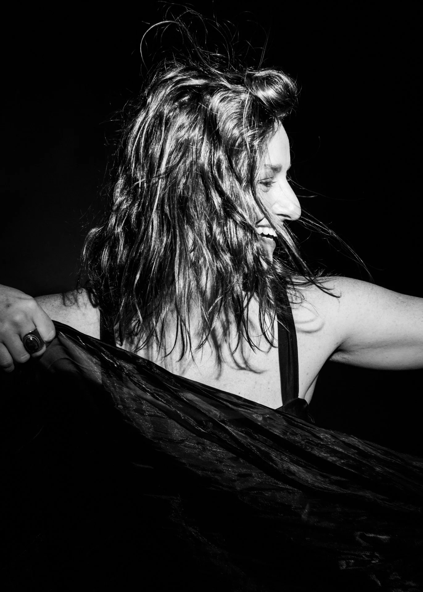 Mujer sonriendo, con cabello rizado y largo, sosteniendo una tela negra, en un fondo oscuro.