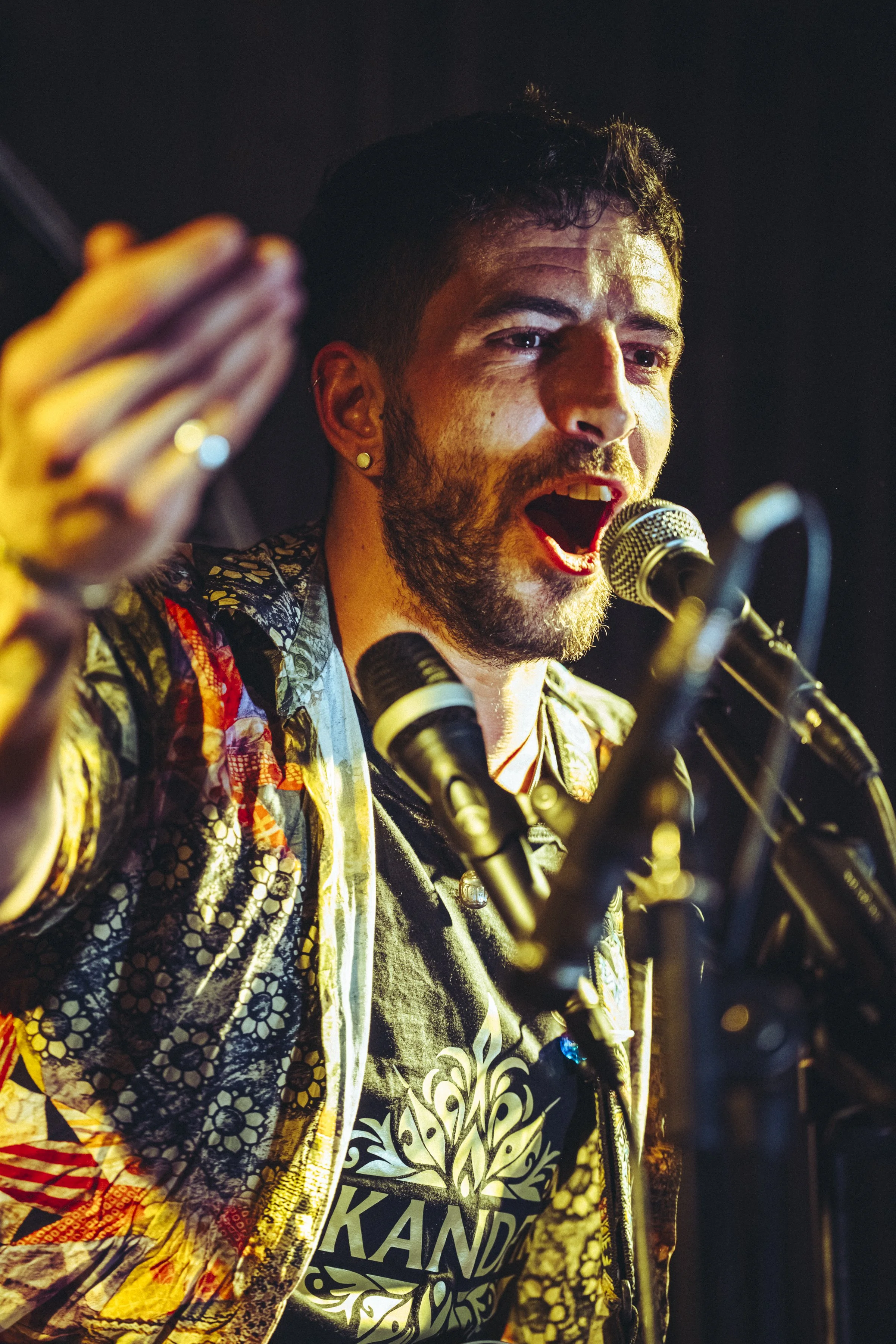 Hombre con barba y tatuajes cantando con micrófono en mano y brazos extendidos, usando camisa con estampados coloridos en un escenario durante una presentación musical.