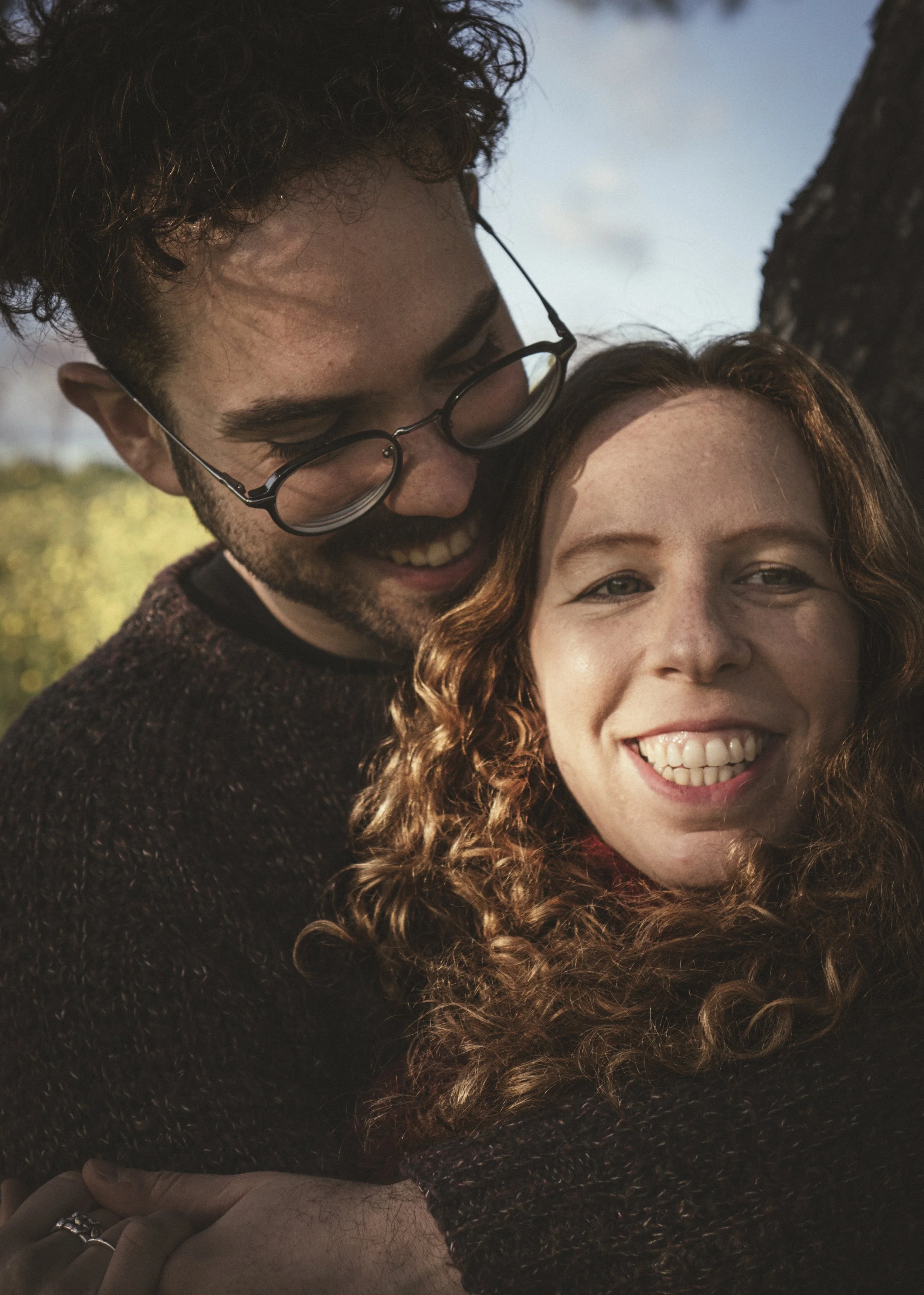 Una pareja sonriendo y abrazándose en un entorno al aire libre, con árbol y cielo de fondo.