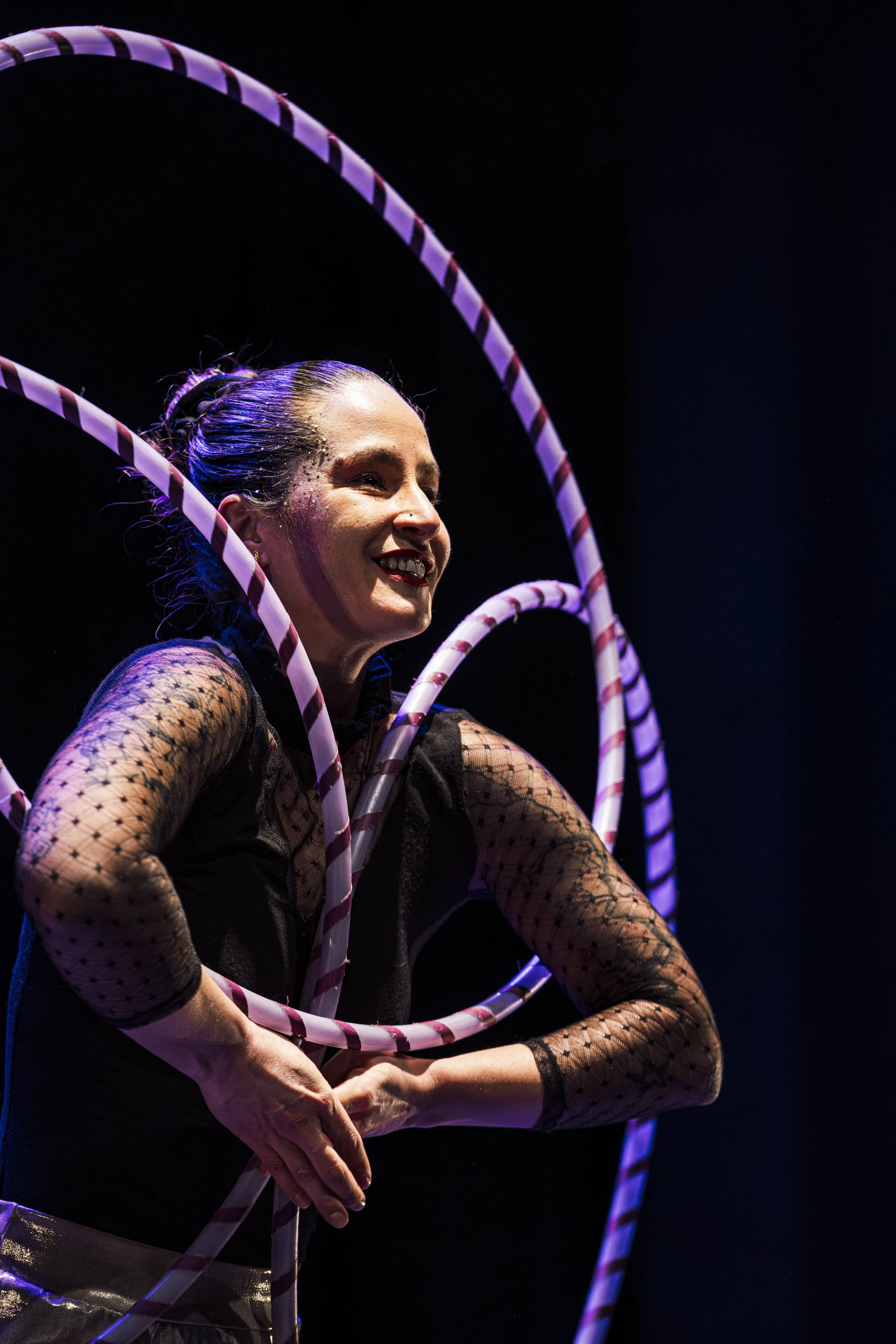 Una mujer sonriente usando vestimenta de circo con varillas de colores y aros de hula hoop en un escenario oscuro.