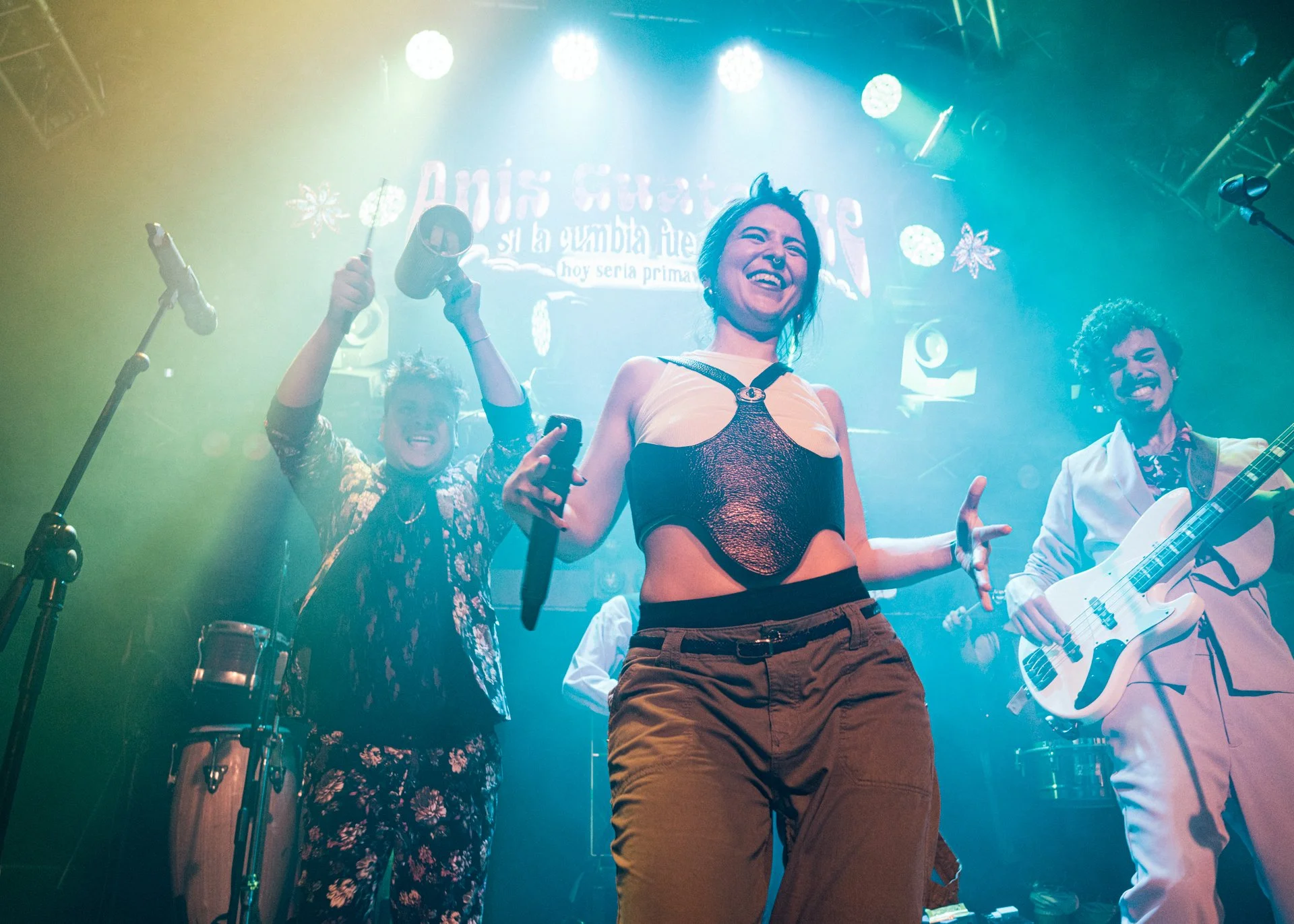 Personas en un escenario de concierto, dos hombres con instrumentos y una mujer en el centro, sonriendo y disfrutando de la música, con luces y decoraciones en el fondo.