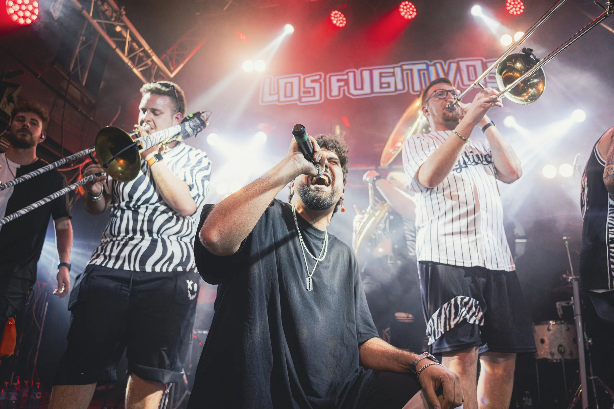 Grupo musical tocando en escenario con luces rojas y blancas, con un cartel que dice 'Los Fugitivos'.
