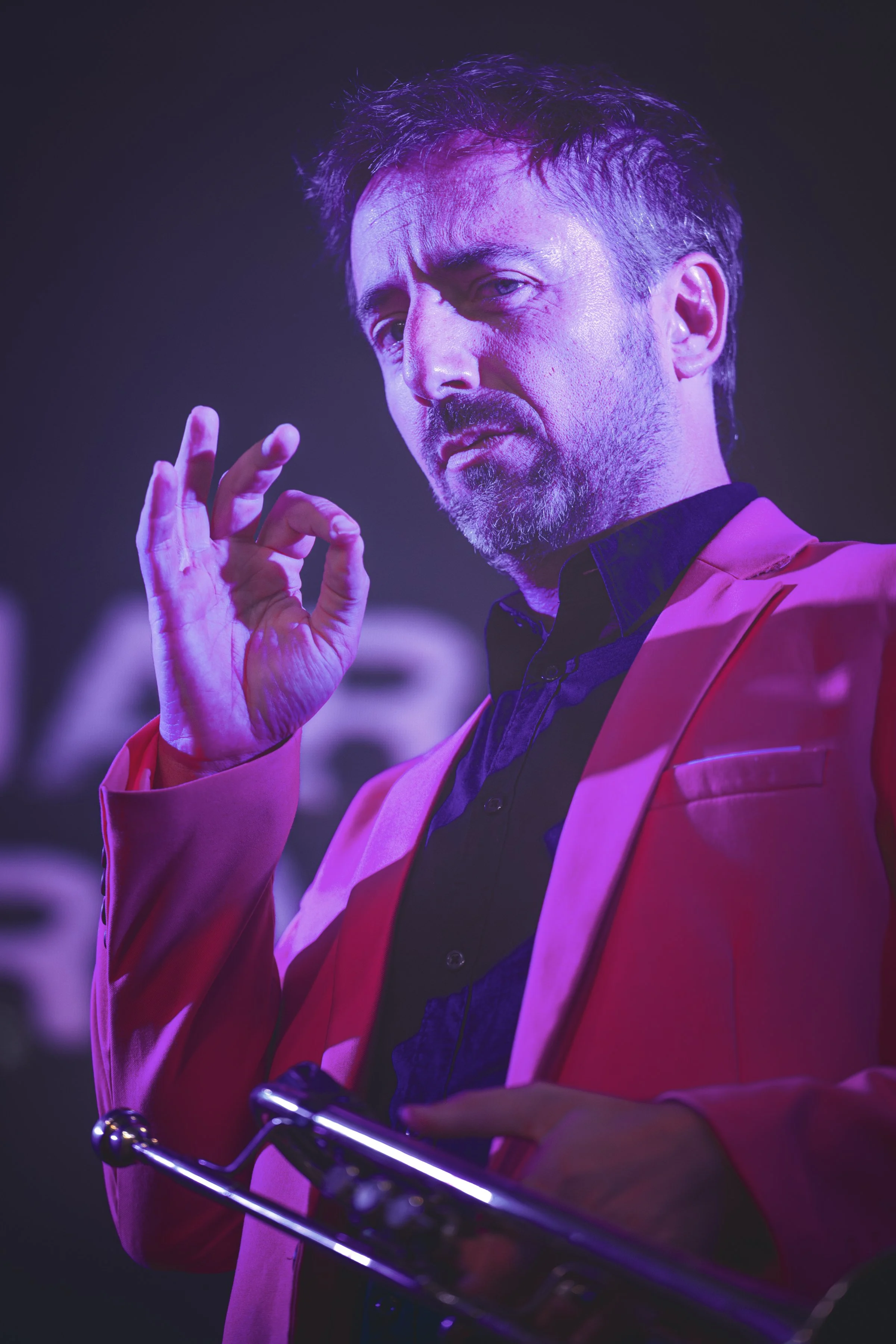 Hombre con barba y cabello oscuro, usando saco rosado y camisa negra, haciendo señal de OK con su mano, en un entorno con iluminación morada y fondo borroso.