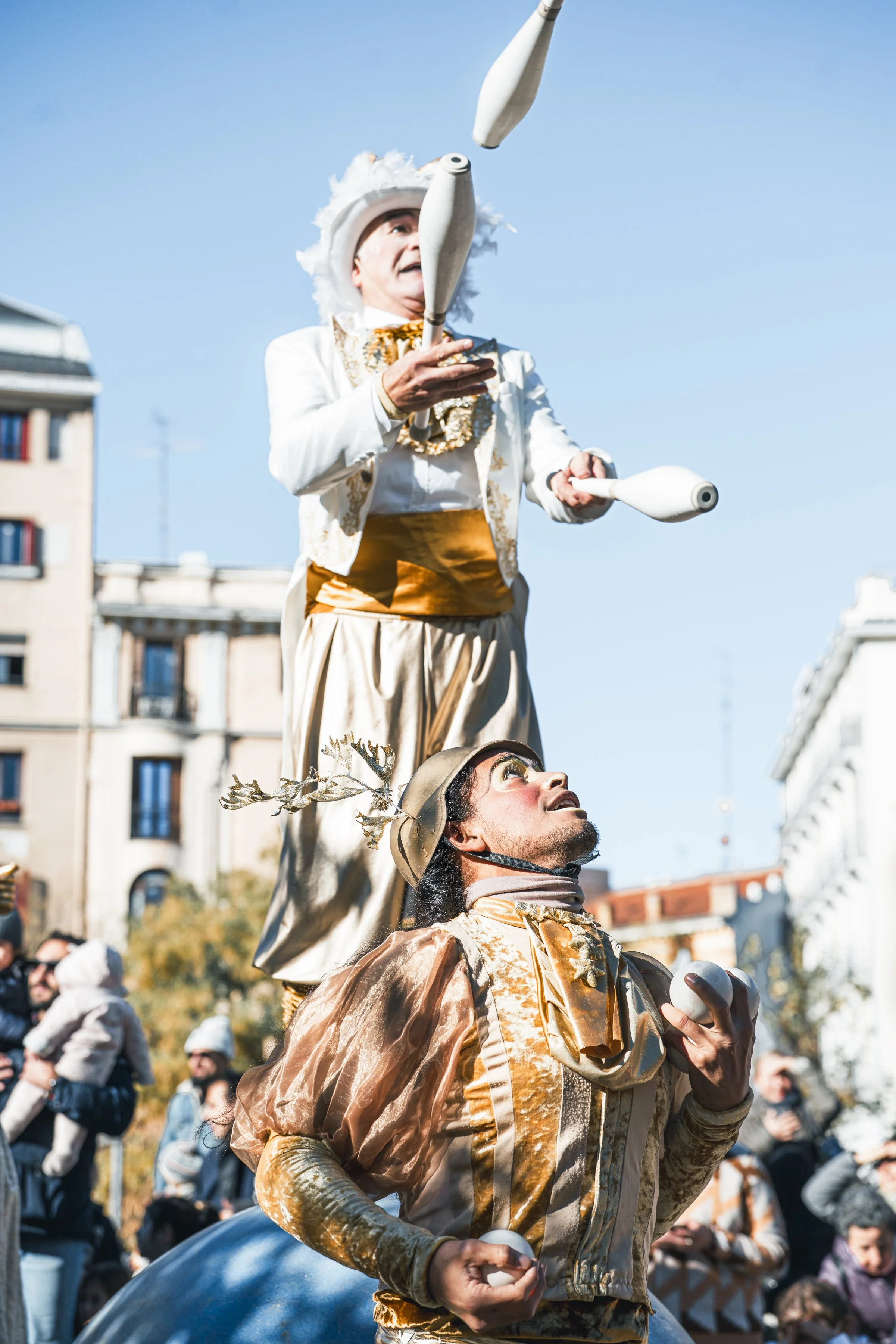 Dos payasos en un desfile, uno de pie y otro en el suelo, haciendo malabares con pelotas en un espacio urbano con edificios y espectadores al fondo.