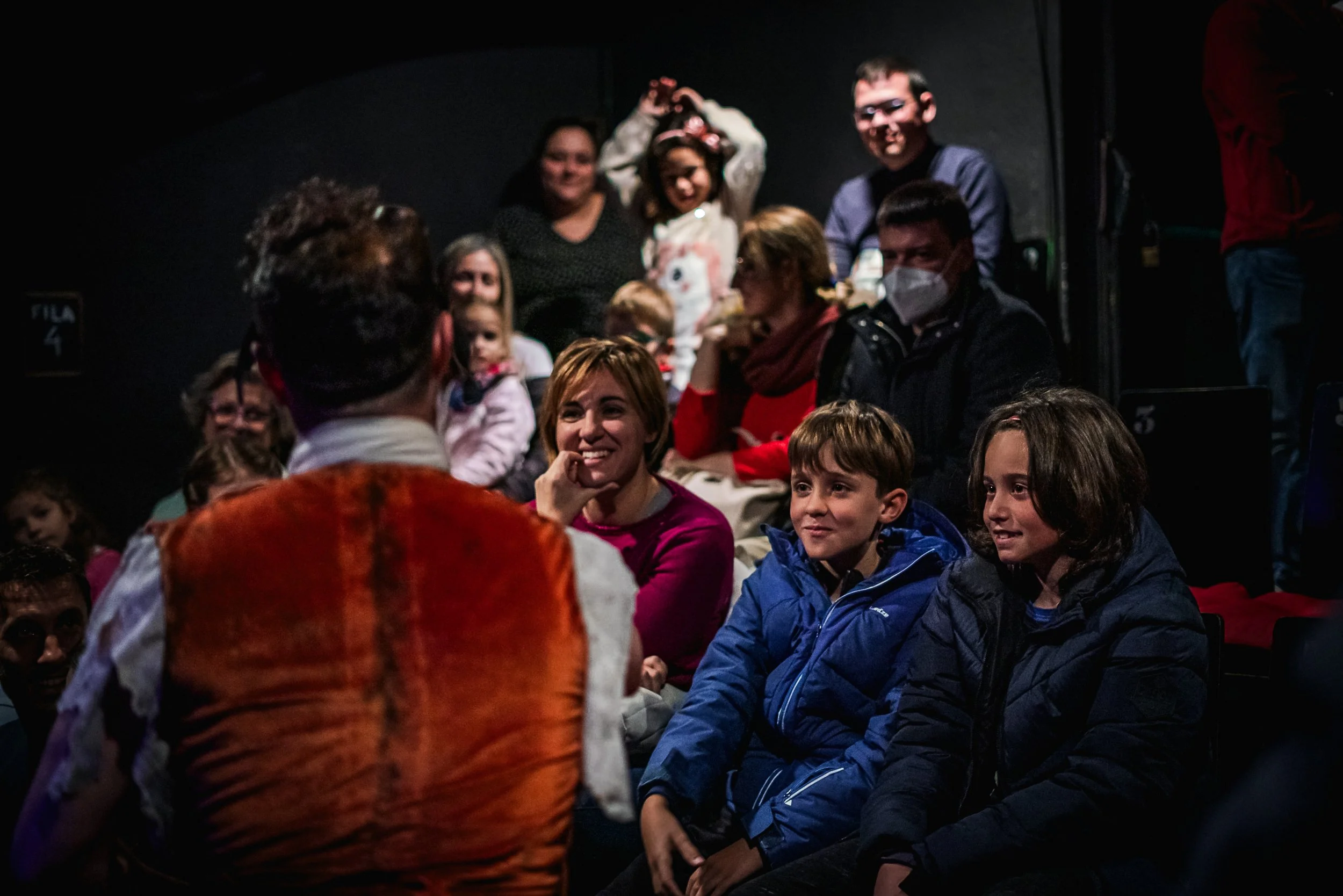 Personas en un teatro interactuando con un actor disfrazado, con niños y adultos en la audiencia, algunos niños sonriendo y un adulto con mascarilla.