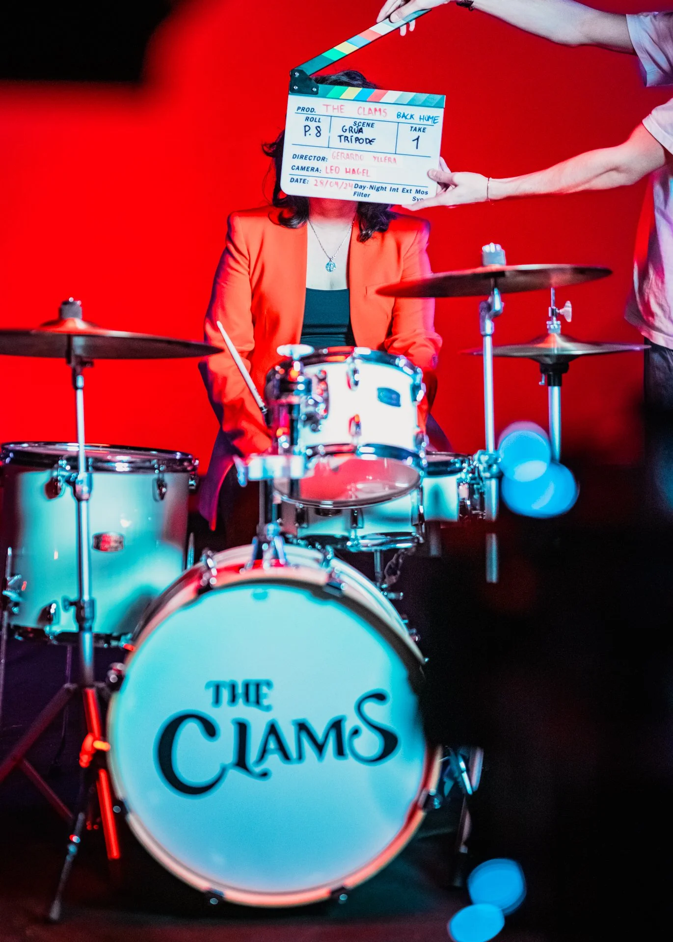 Una mujer con chaqueta roja sentada detrás de una batería con el logotipo de 'The Clams'. Una persona al lado sostiene una claqueta de rodaje frente a su rostro, en un escenario con fondo rojo.