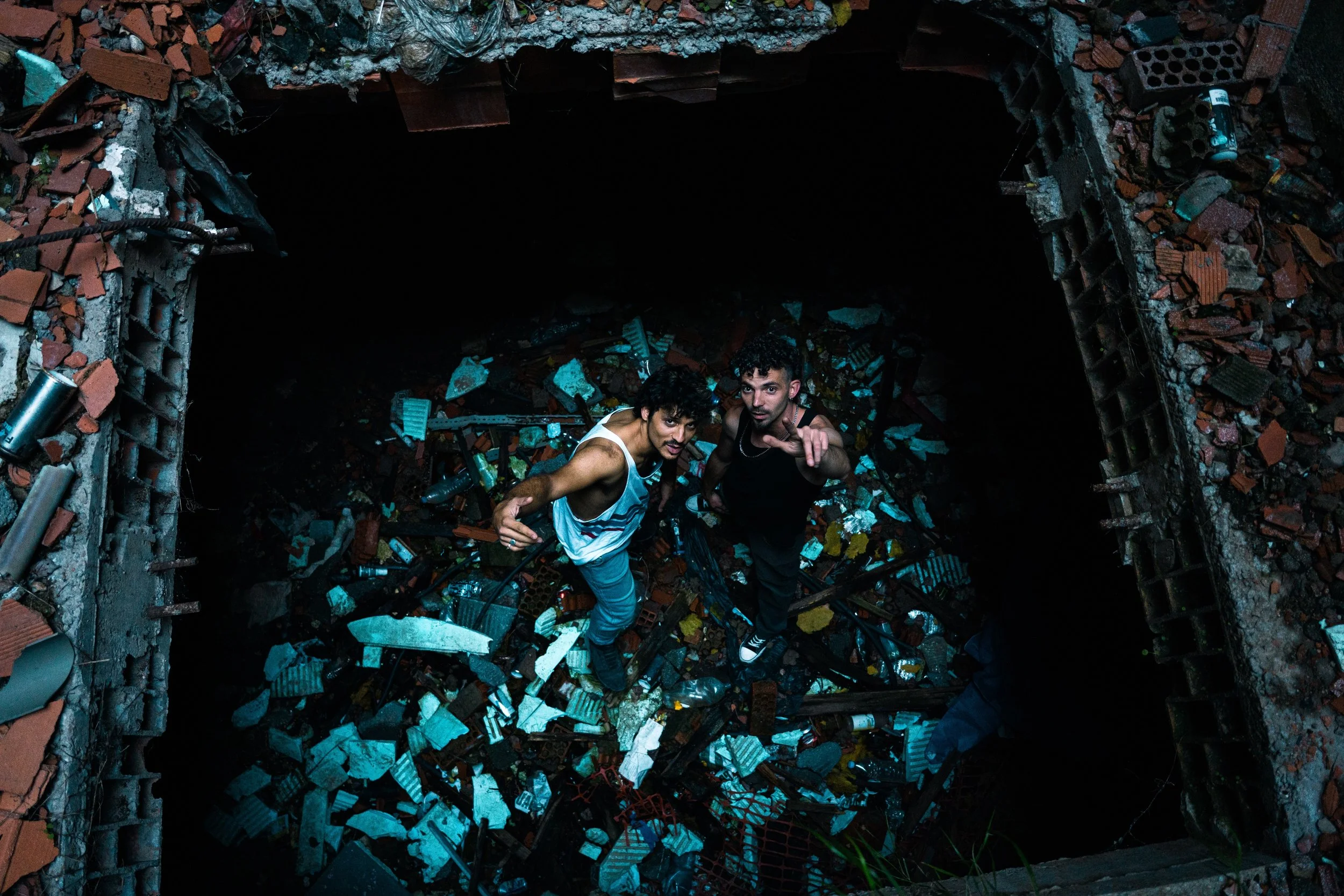 Dos personas de pie sobre basura en un escombrera, mirando hacia arriba, en un entorno oscuro y desordenado.