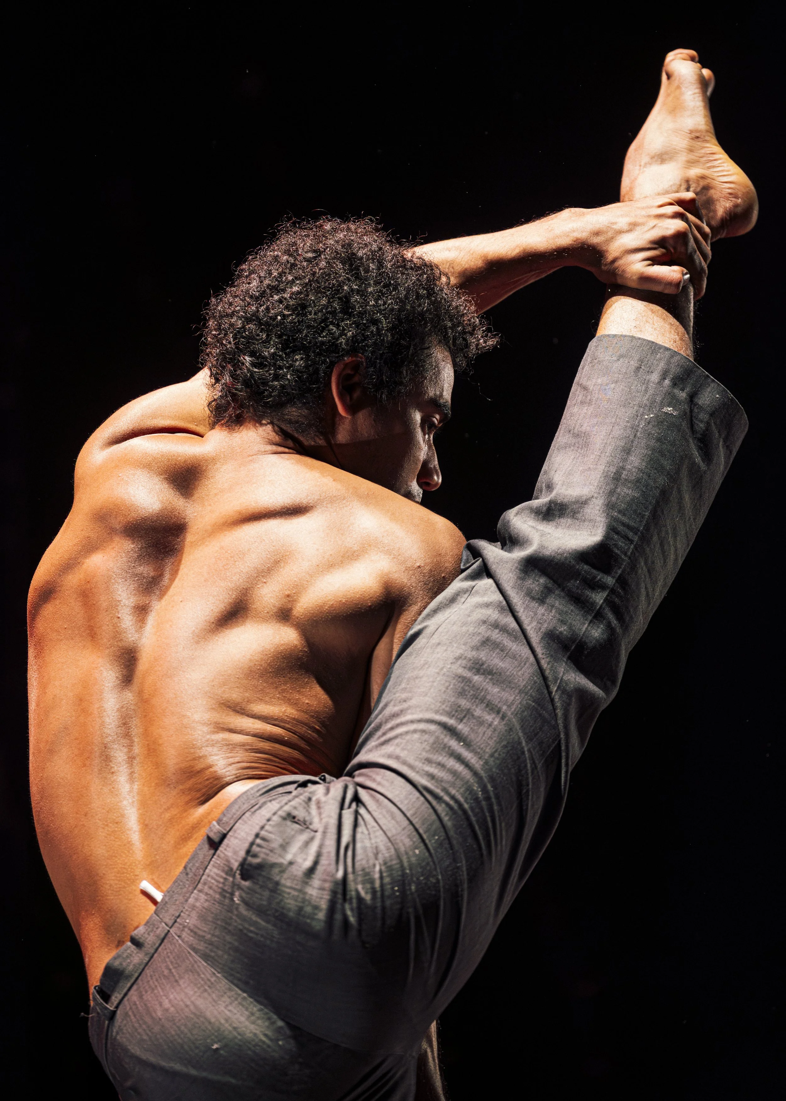 Hombre sin camisa realizando una postura de estiramiento o ejercicio de equilibrio, con fondo negro.