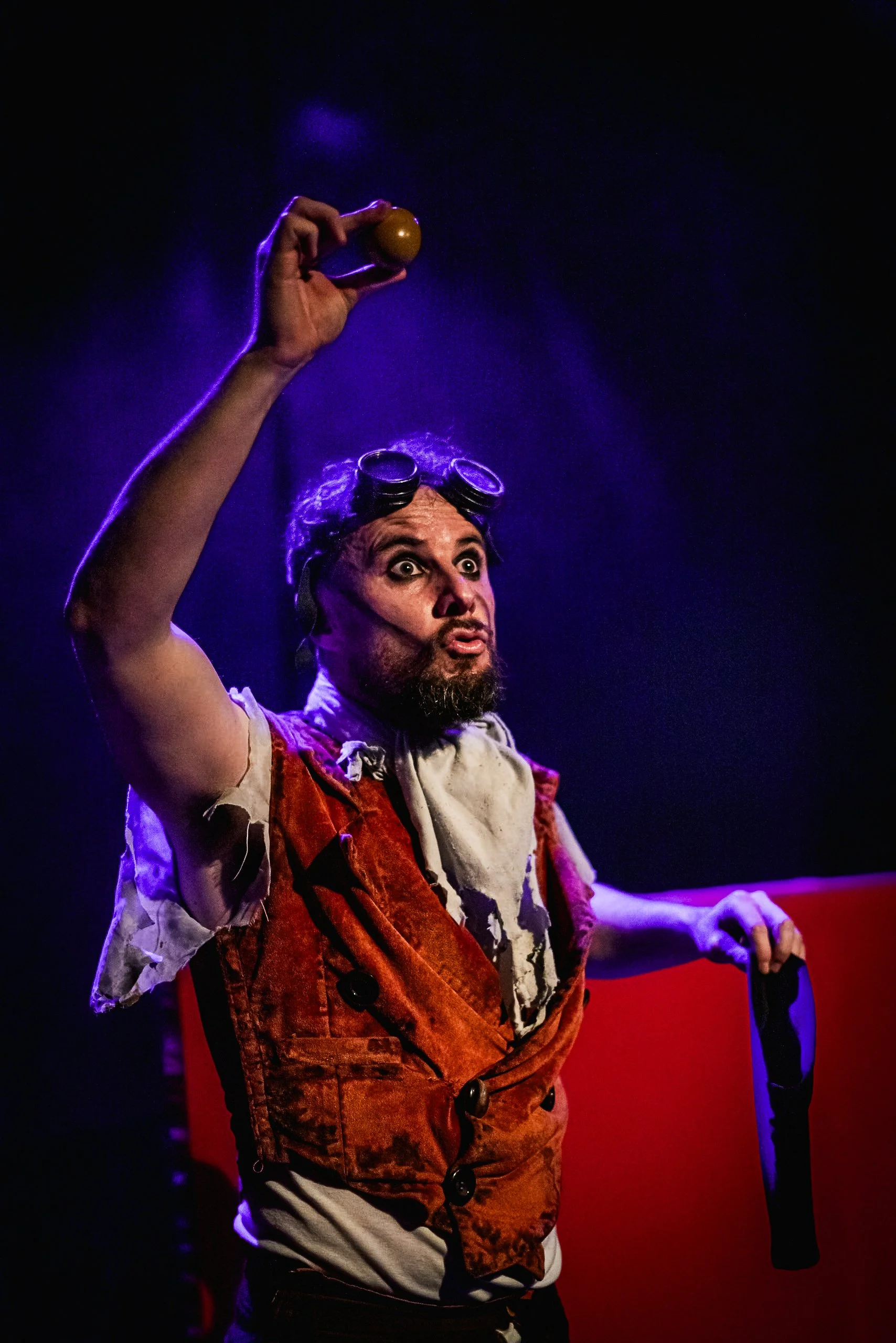 Un hombre con gafas y barba interpretando un personaje teatral, sosteniendo una pelota amarilla en una mano y un objeto negro en la otra, en un escenario con iluminación azul y roja.