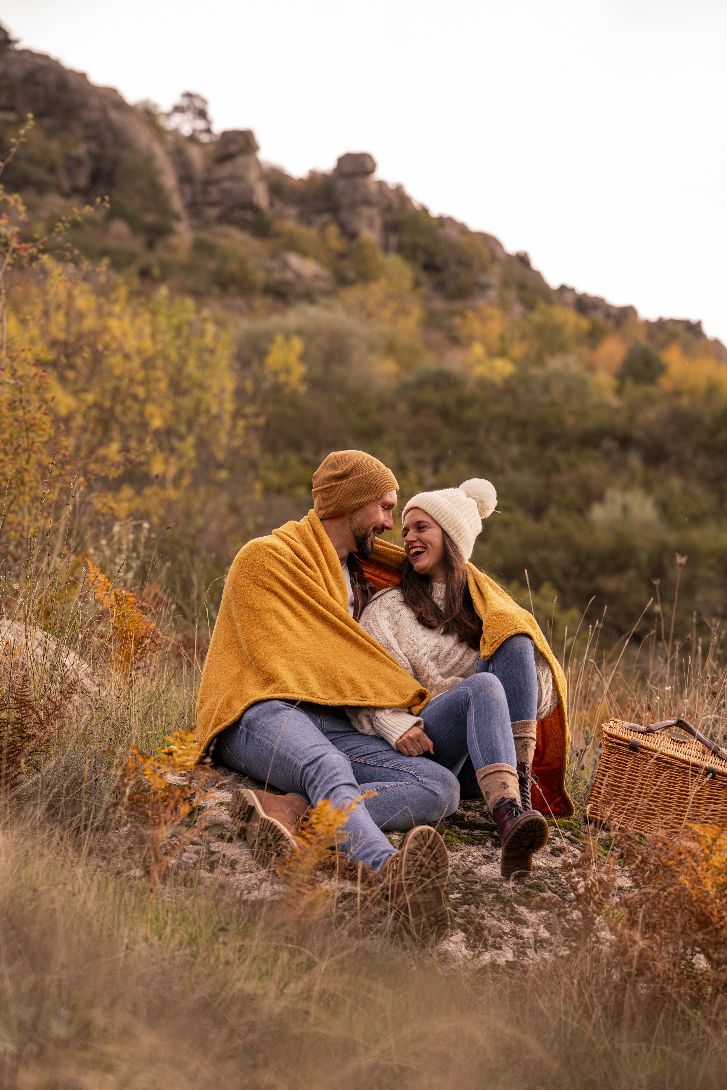 Una pareja sentada en una roca rodeados de vegetación otoñal, compartiendo un momento alegre con un picnic y cubiertos con un cobertor amarillo, en un paisaje de montañas con árboles en colores de otoño.