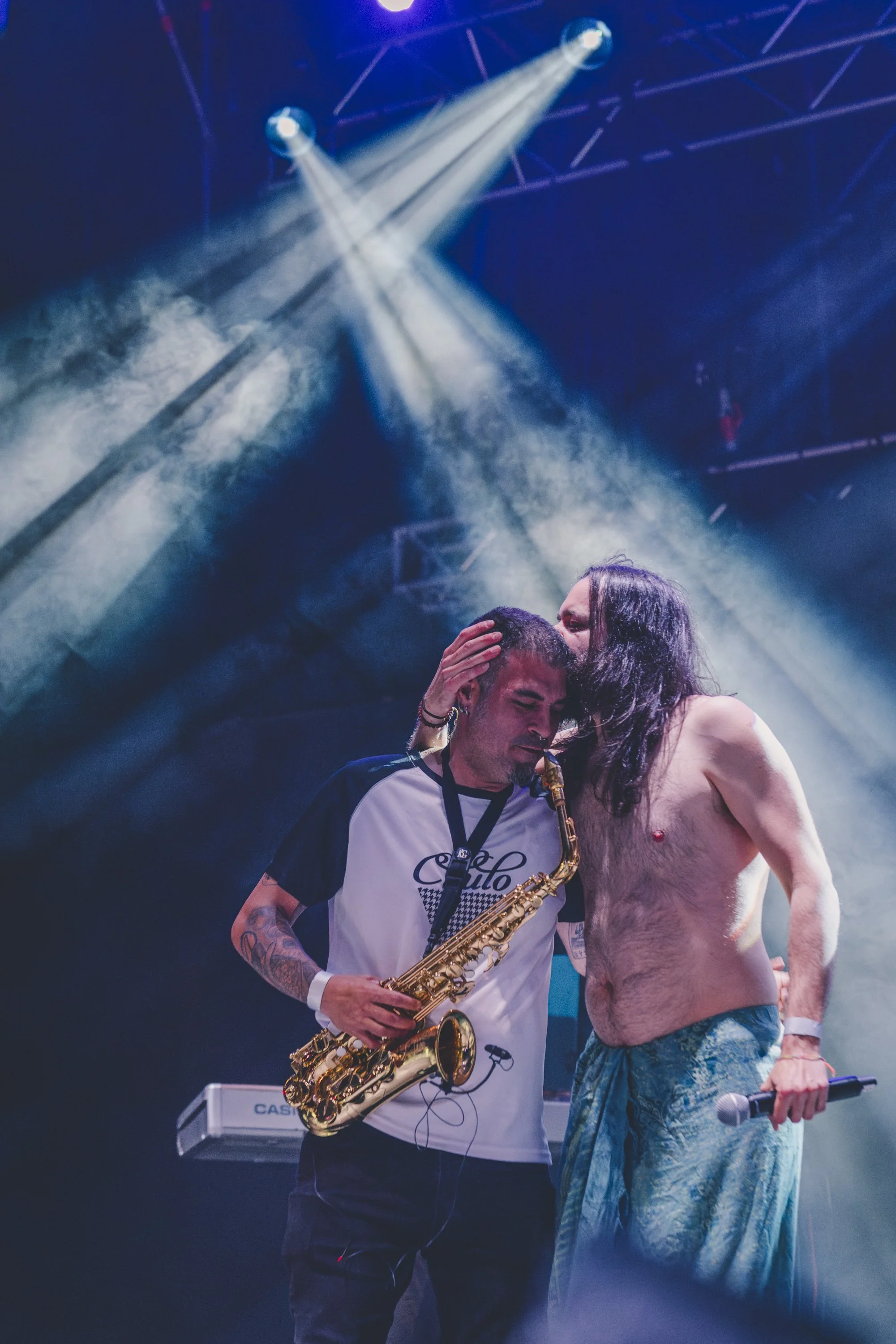 Dos hombres en un escenario, uno tocando el saxofón y otro sin camisa, juntos en un momento emotivo con luces y humo.