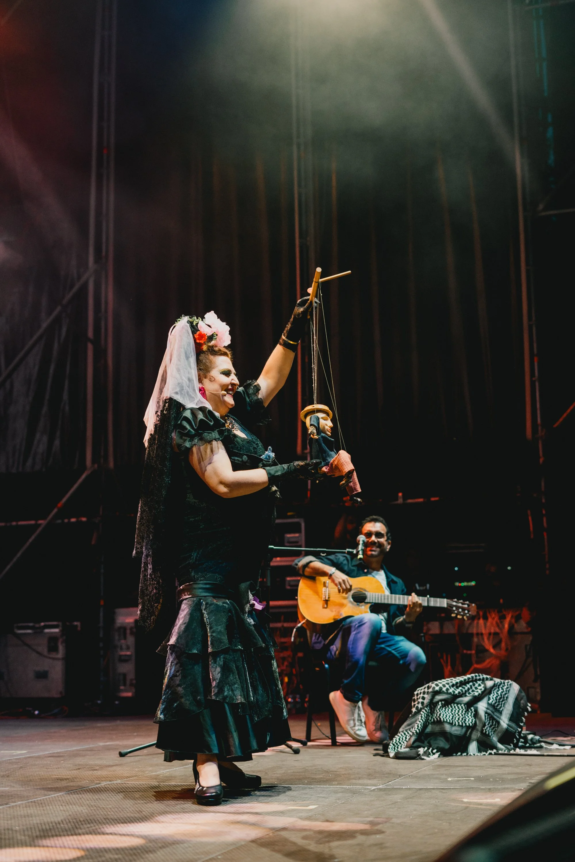 Una mujer con vestimenta de estilo tradicional y accesorios de dia de muertos, realizando una actuación con marionetas en un escenario con un guitarrista acompañándola, en un ambiente de concierto o teatro.