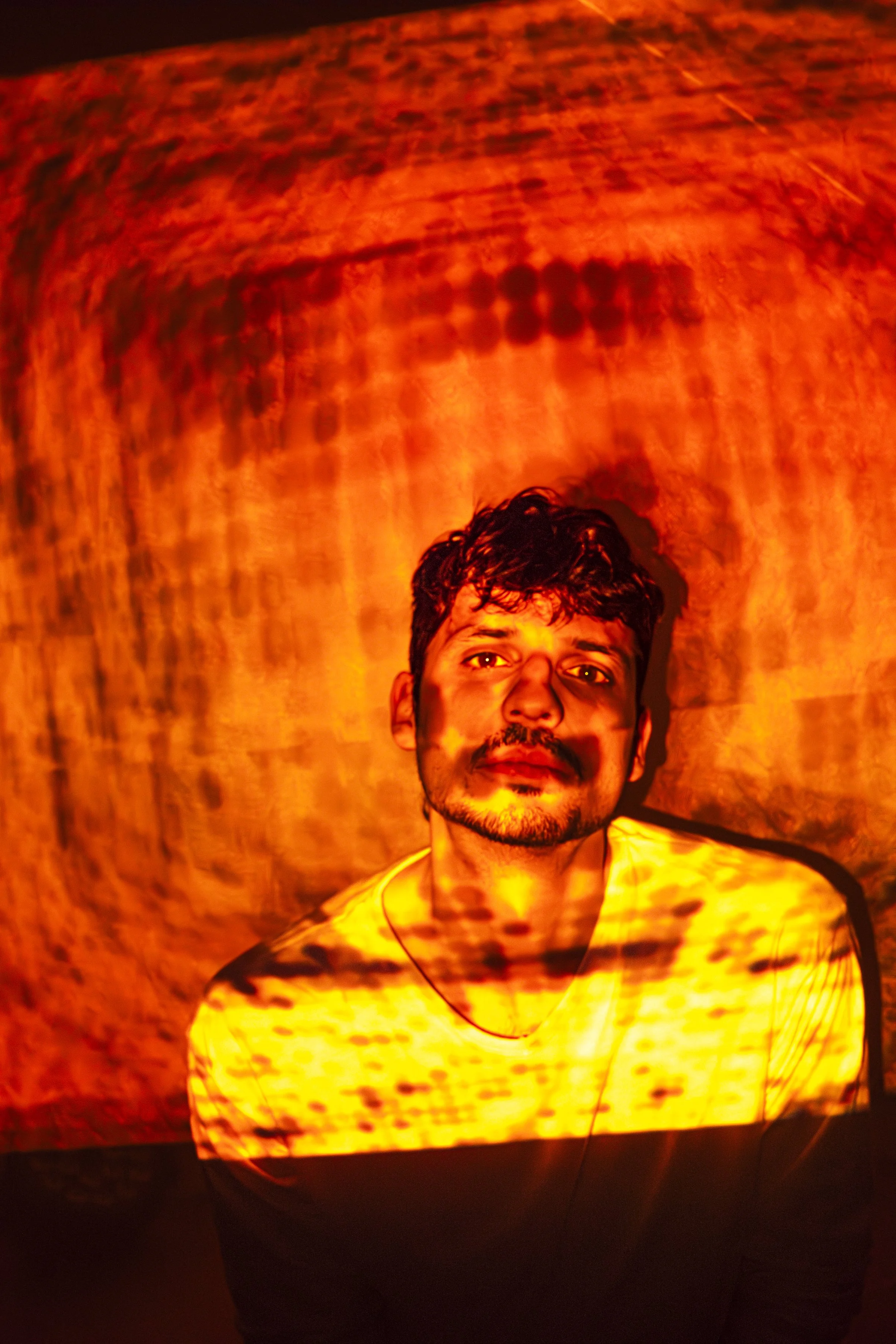 Hombre con barba y cabello rizado, iluminado por una luz anaranjada que crea patrones en su rostro y fondo, con expresión neutral.