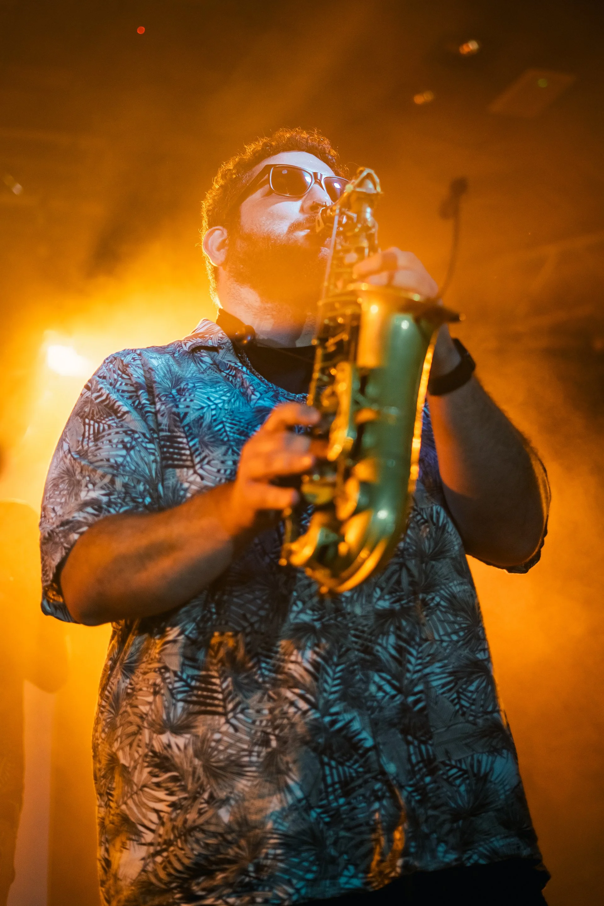 Hombre tocando el saxofón en un escenario con iluminación naranja y con gafas de sol.