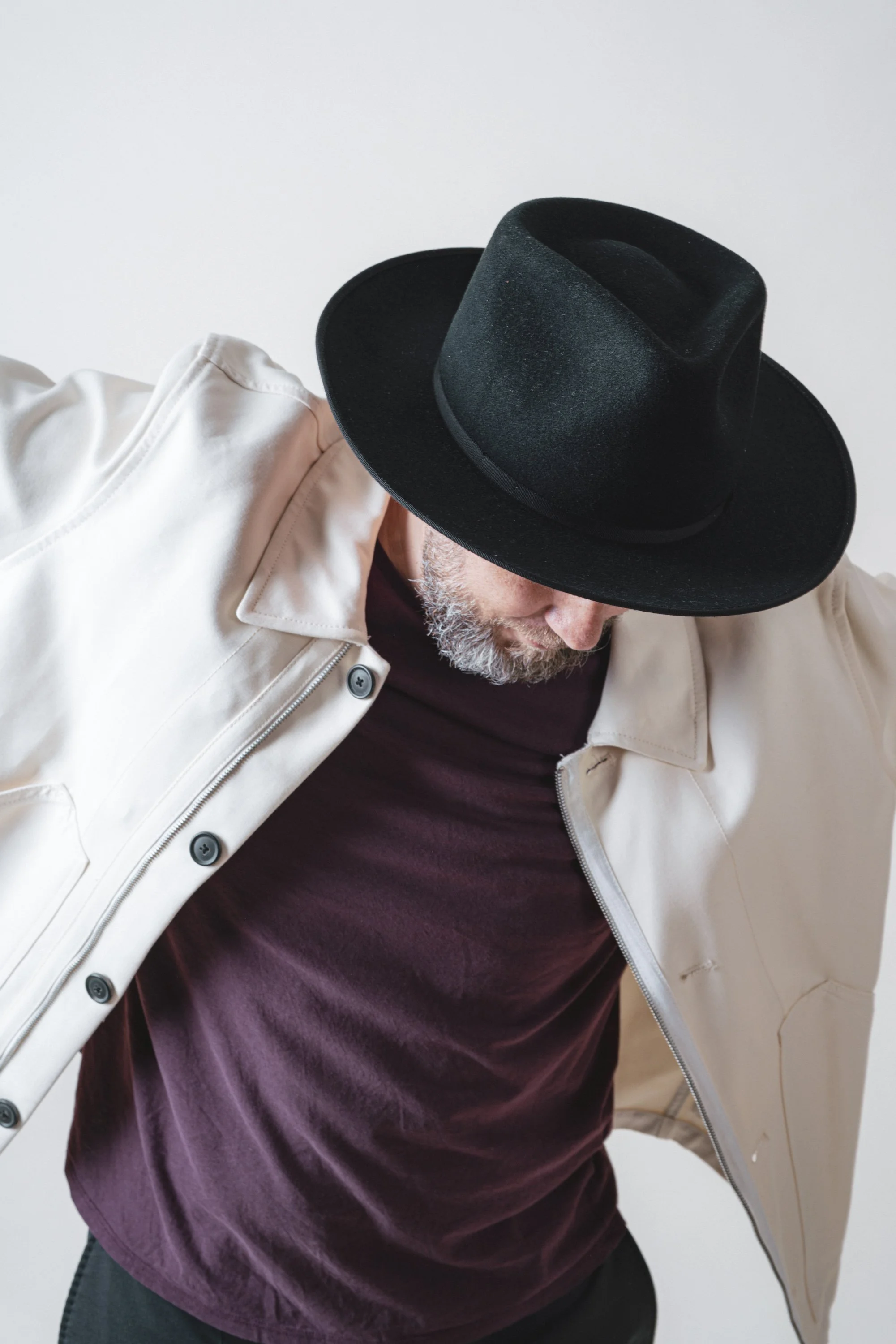 Hombre con gorra negra, camiseta morada y chaqueta blanca en posición inclinada hacia abajo.