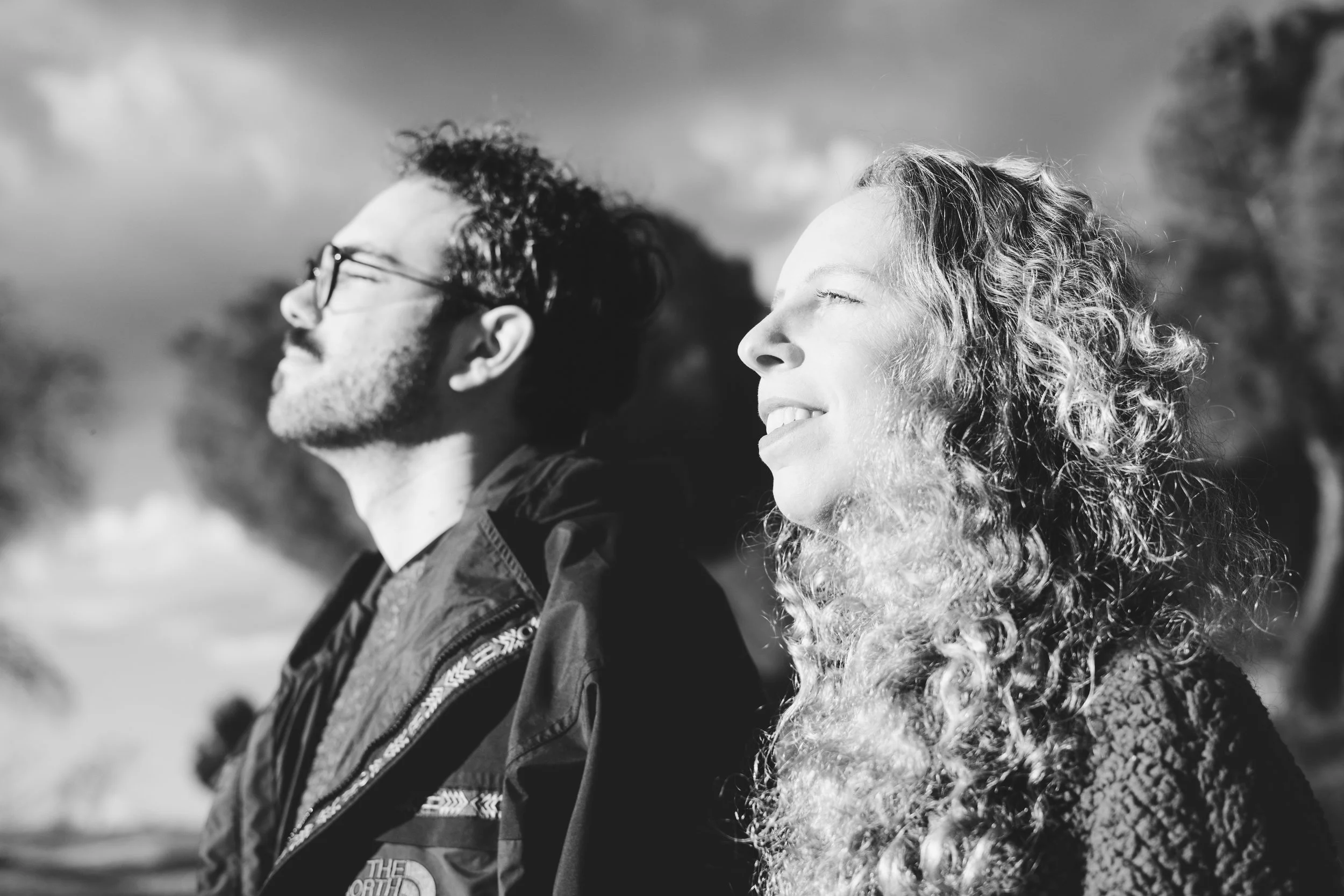 Dos personas de espaldas, una mujer sonriendo con ojos cerrados y un hombre con gafas y barba, disfrutando del sol al aire libre.
