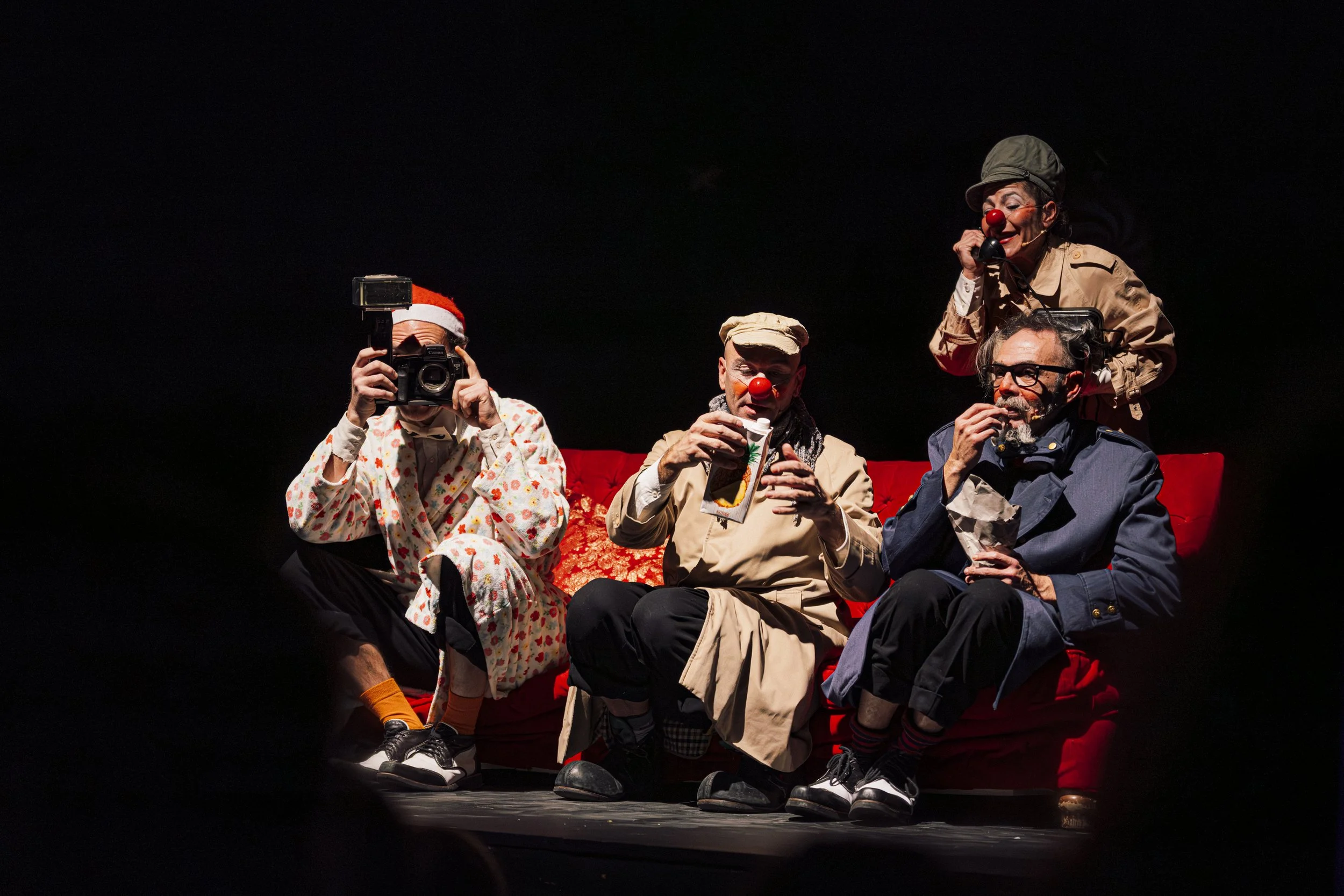 Cuatro payasos en un escenario con fondo negro, uno con disfraz de Navidad, otro con trajes casuales y dos con accesorios de payaso, una de ellas hablando por teléfono y la otra tomando una bebida, todos en una escena de comedia.