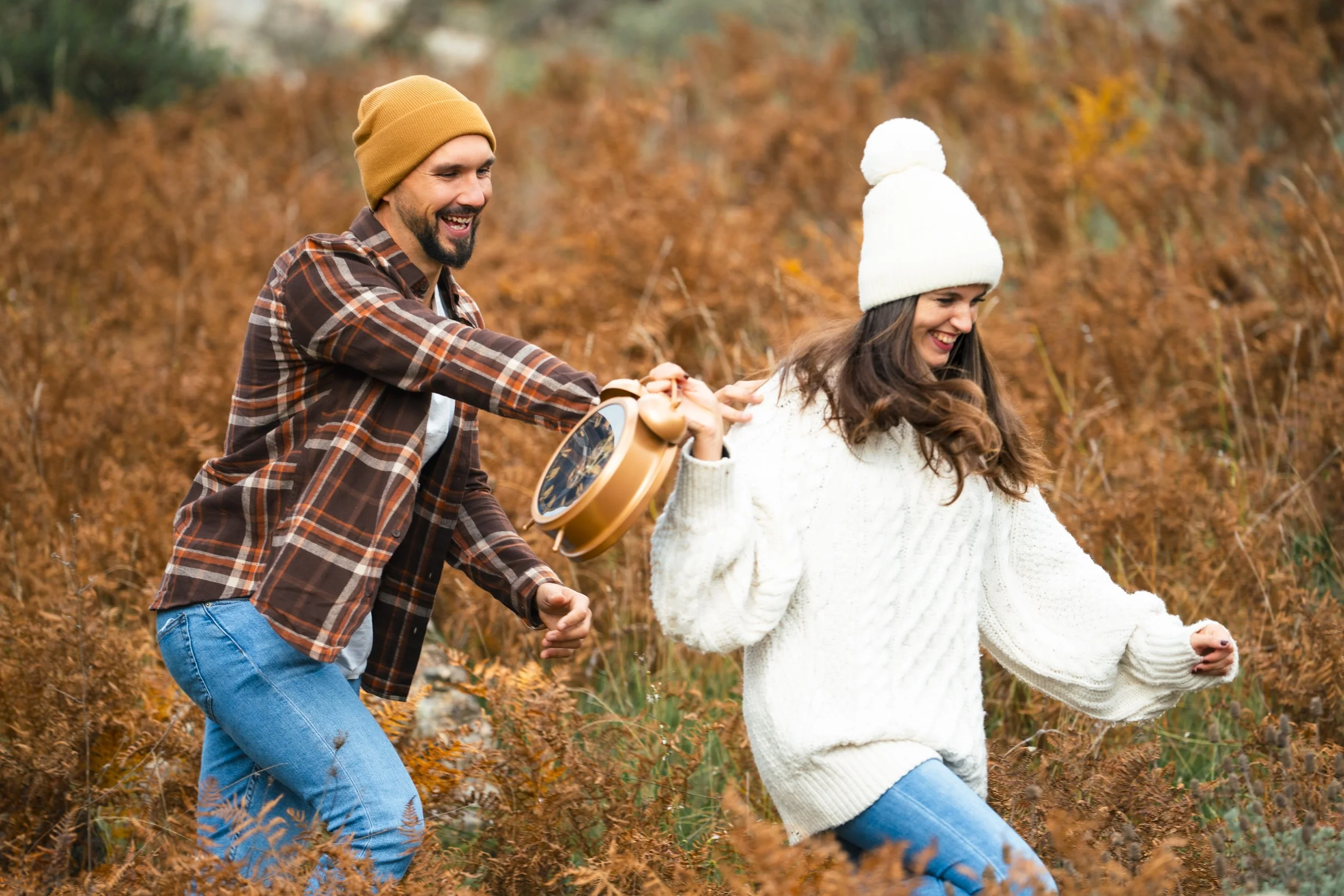 Una pareja feliz en un campo de otoño con hojas secas, la mujer con gorro blanco y suéter blanco y el hombre con gorro color mostaza y chaqueta de cuadros, ambos disfrutando del día.
