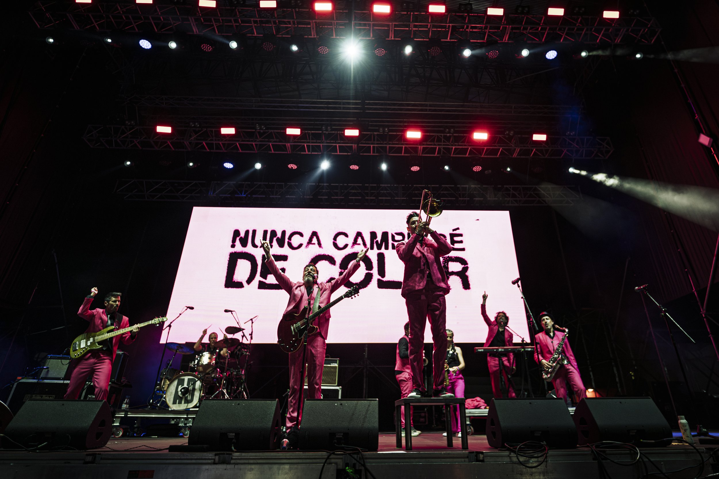 Grupo de músicos en un concierto en vivo, tocando instrumentos en un escenario con fondo digital que dice 'Nunca cambiaré de color', con luces de escenario y equipo de sonido.