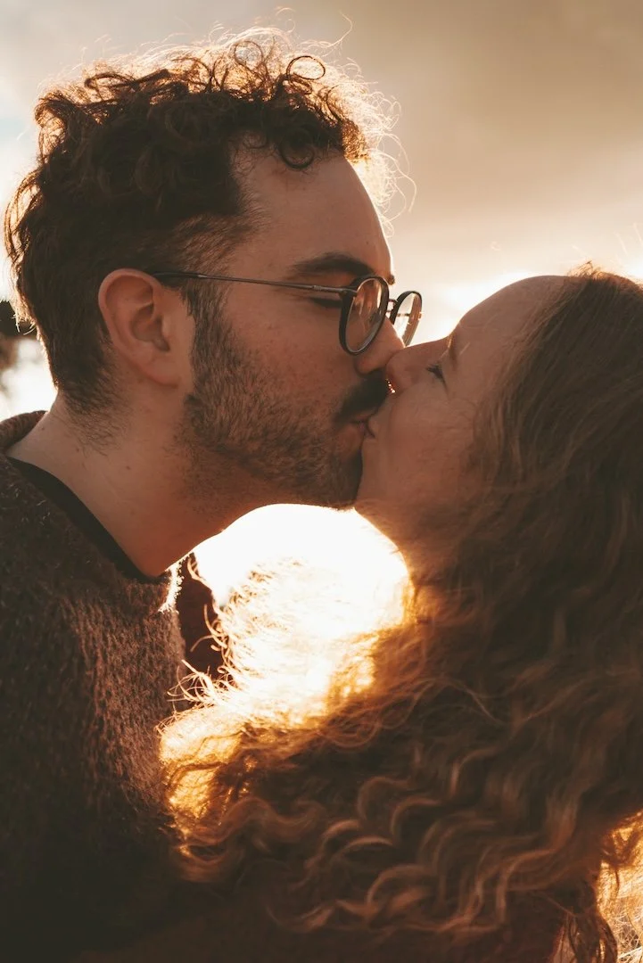 Una pareja se besa al atardecer, con luz cálida y nubes en el cielo.