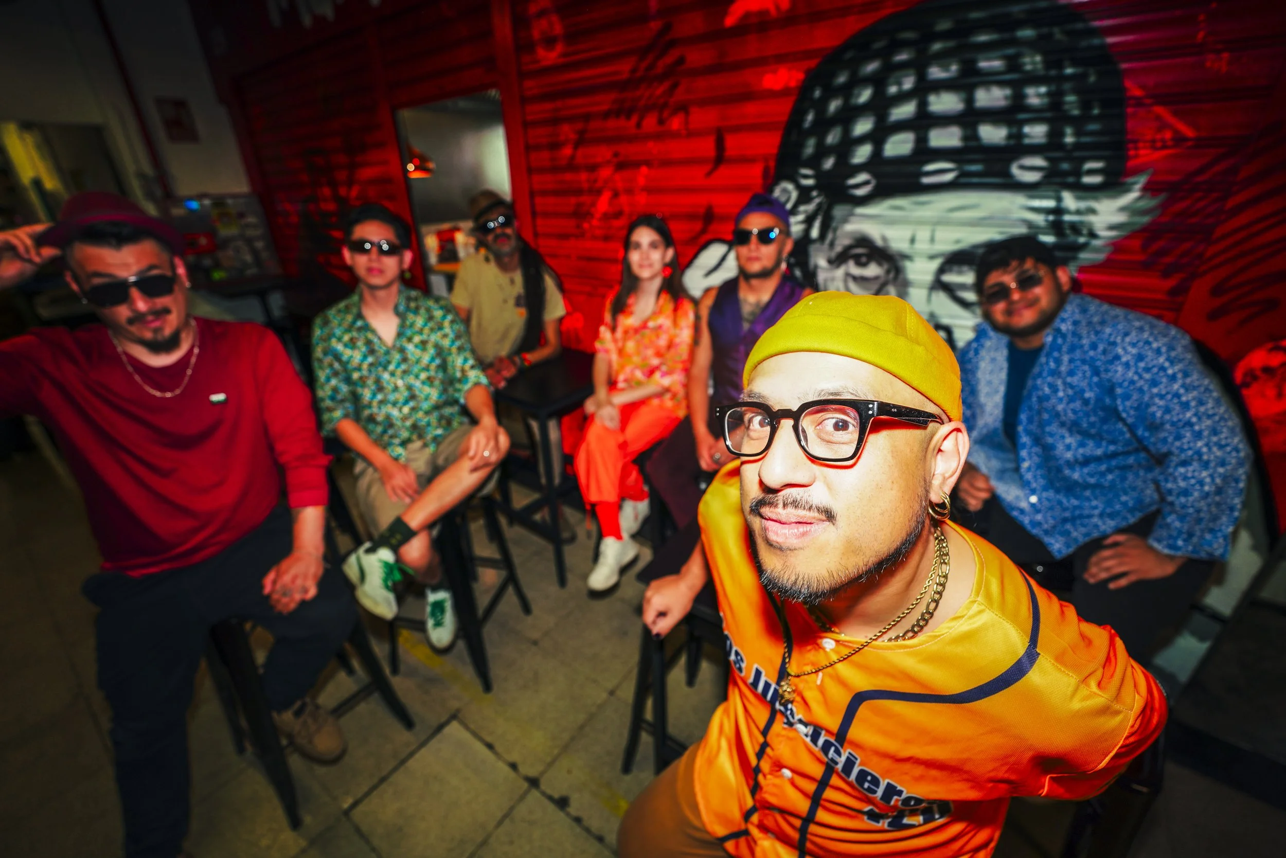 Grupo de personas en un lugar con paredes de madera, algunos usando gafas oscuras, ropa colorida y accesorios llamativos, uno en primer plano lleva gafas grandes negras y una gorra amarilla, todos parecen estar en un evento social o fiesta.