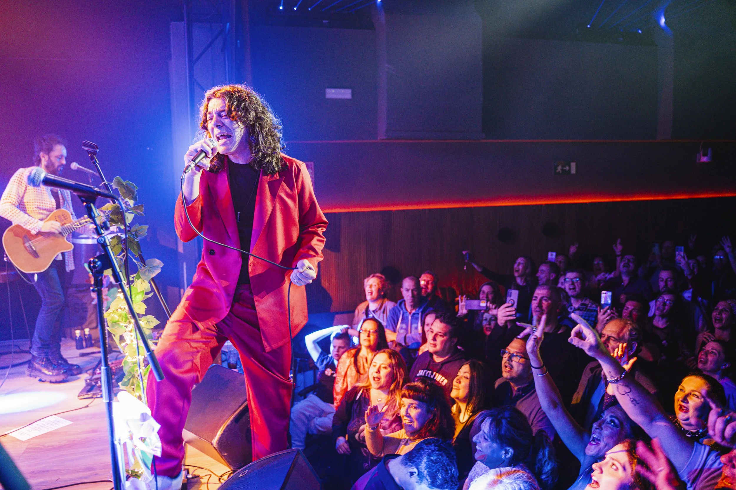 Una cantante con un abrigo rojo en concierto, acompañada de un guitarrista, frente a un público emocionado que toma fotos y videoclips con sus teléfonos.