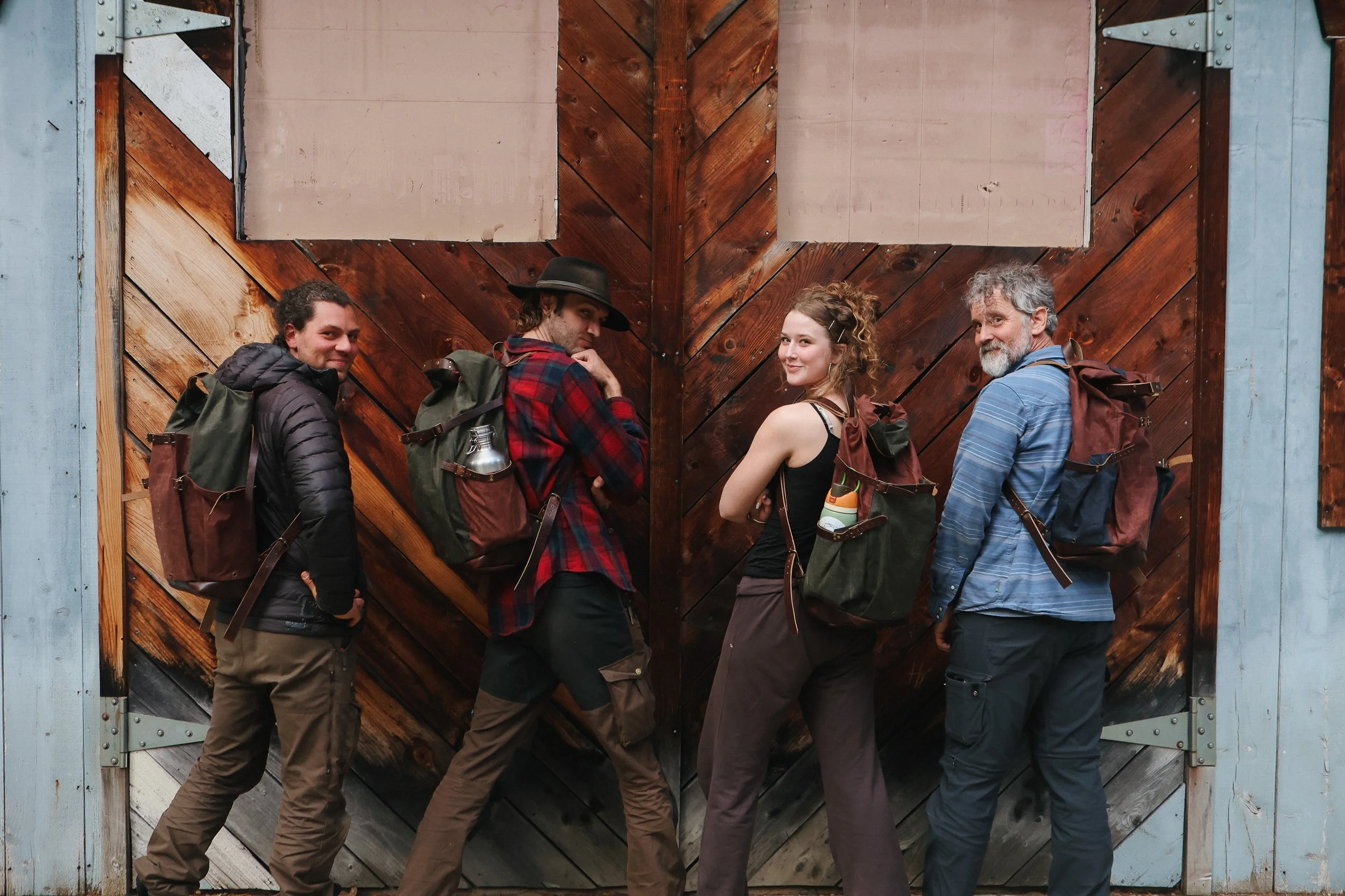 Four people standing in a row outdoors, leaning against a large, wooden, cross-braced barn door wearing hand made backpacks and smiling.