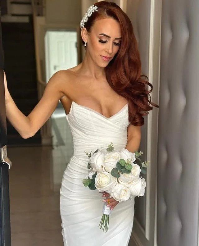 A bride in a strapless white wedding dress holding a bouquet of white roses and greenery, standing indoors with a gentle smile, her long red hair styled in loose curls, wearing a floral headpiece and earrings.